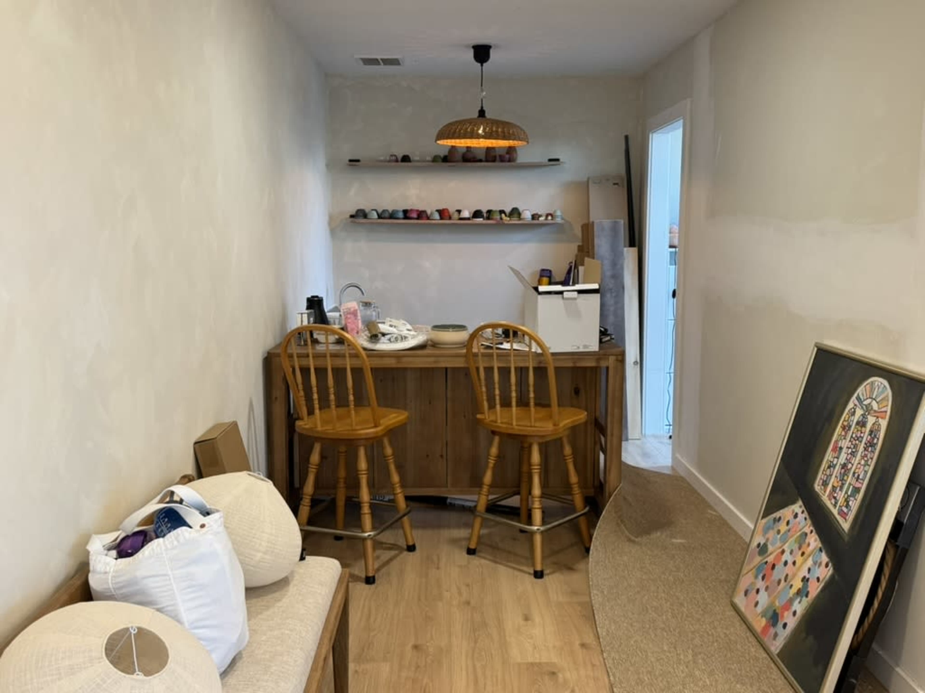 The image shows a small room with two wooden barstools at a counter, a few objects on a shelf above, and a large framed painting on the floor.
