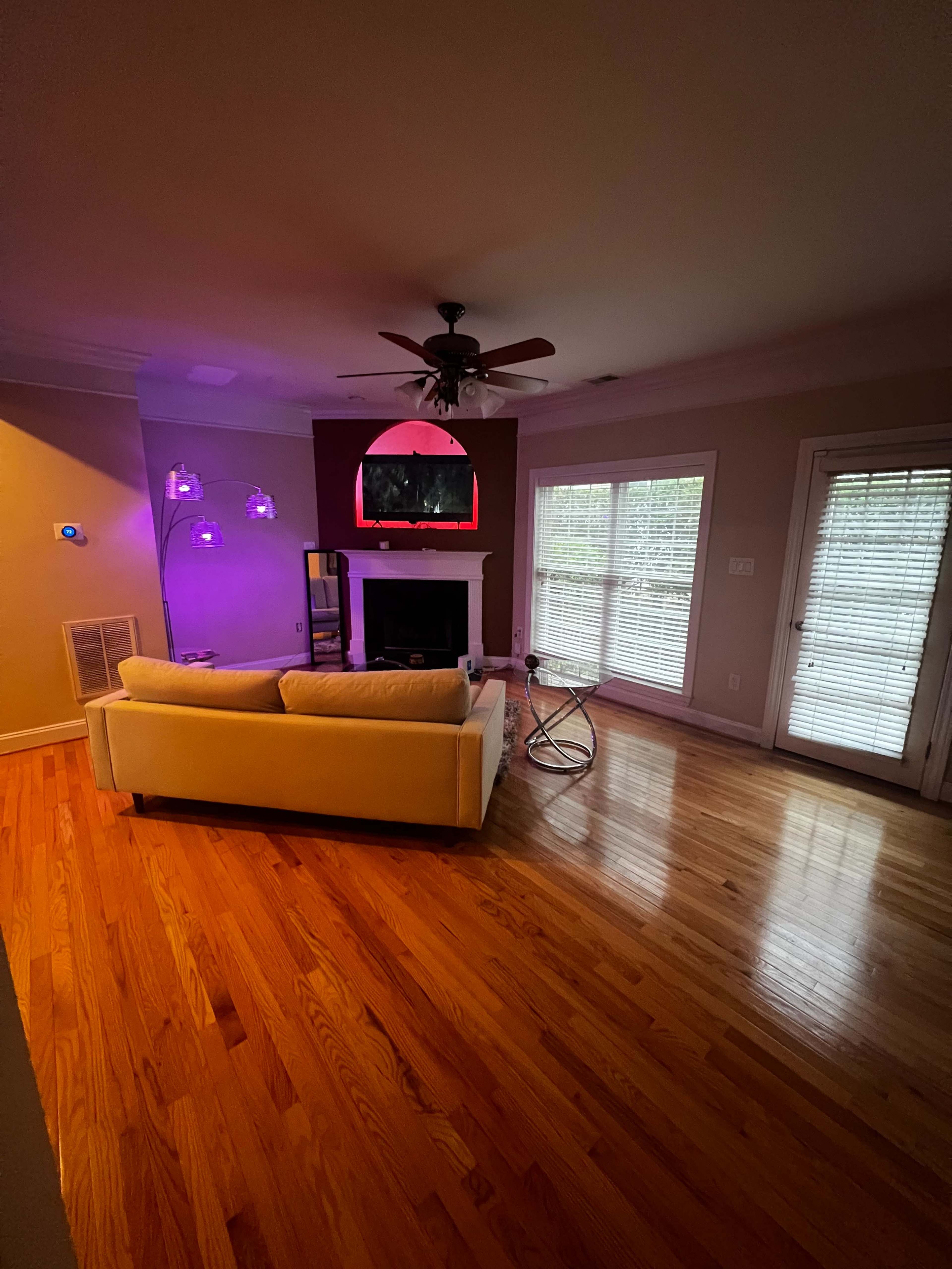 The living room features a beige sofa, a fireplace, a mounted TV, and large windows with blinds, all set on polished wooden flooring.