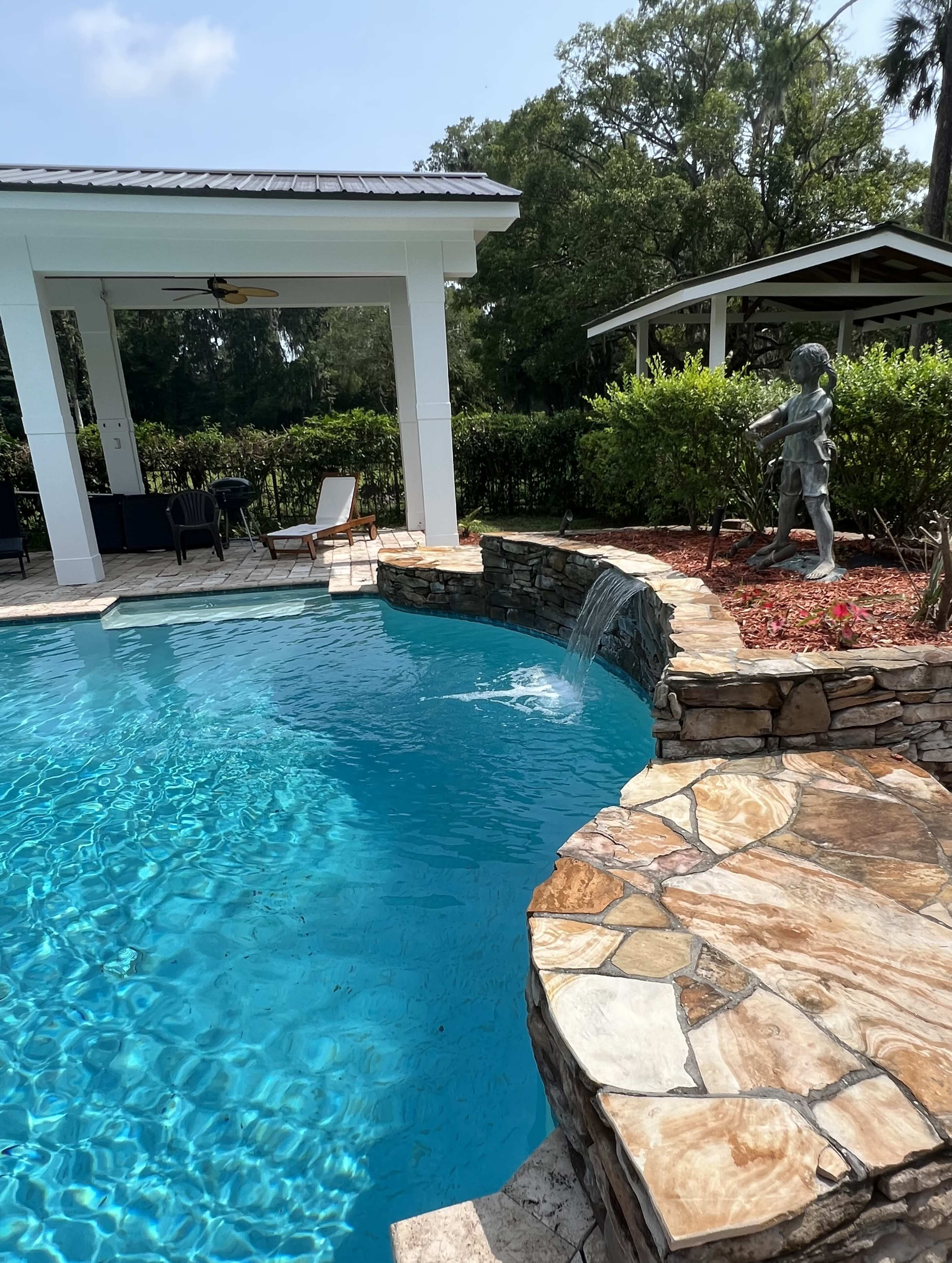 A stone pool deck surrounds a rectangular swimming pool that features a waterfall cascading from a rock ledge, with a sculpture of a figure nearby.