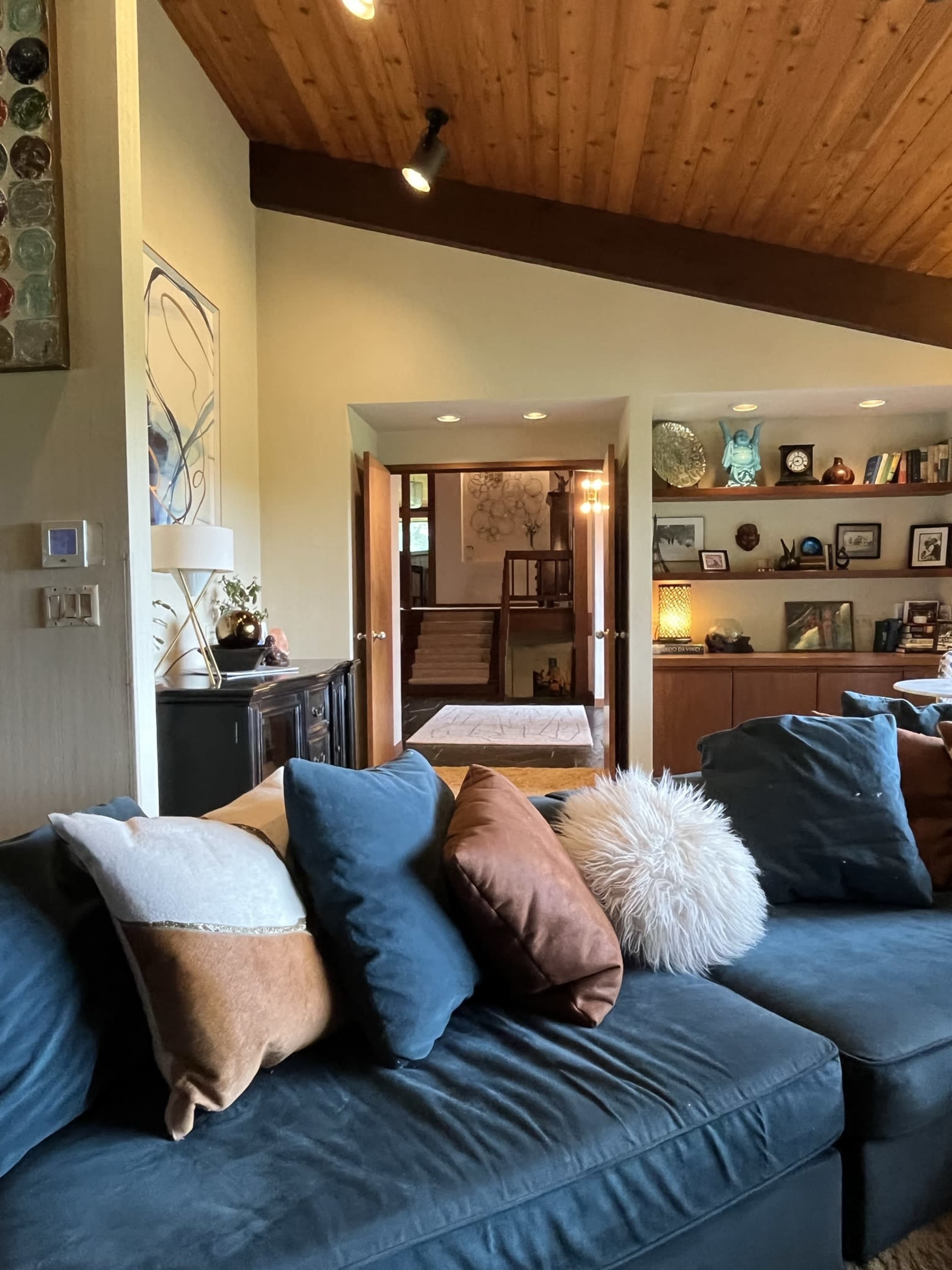 A living room features a blue couch adorned with decorative pillows and a view into a hallway leading to a staircase and an adjoining room.