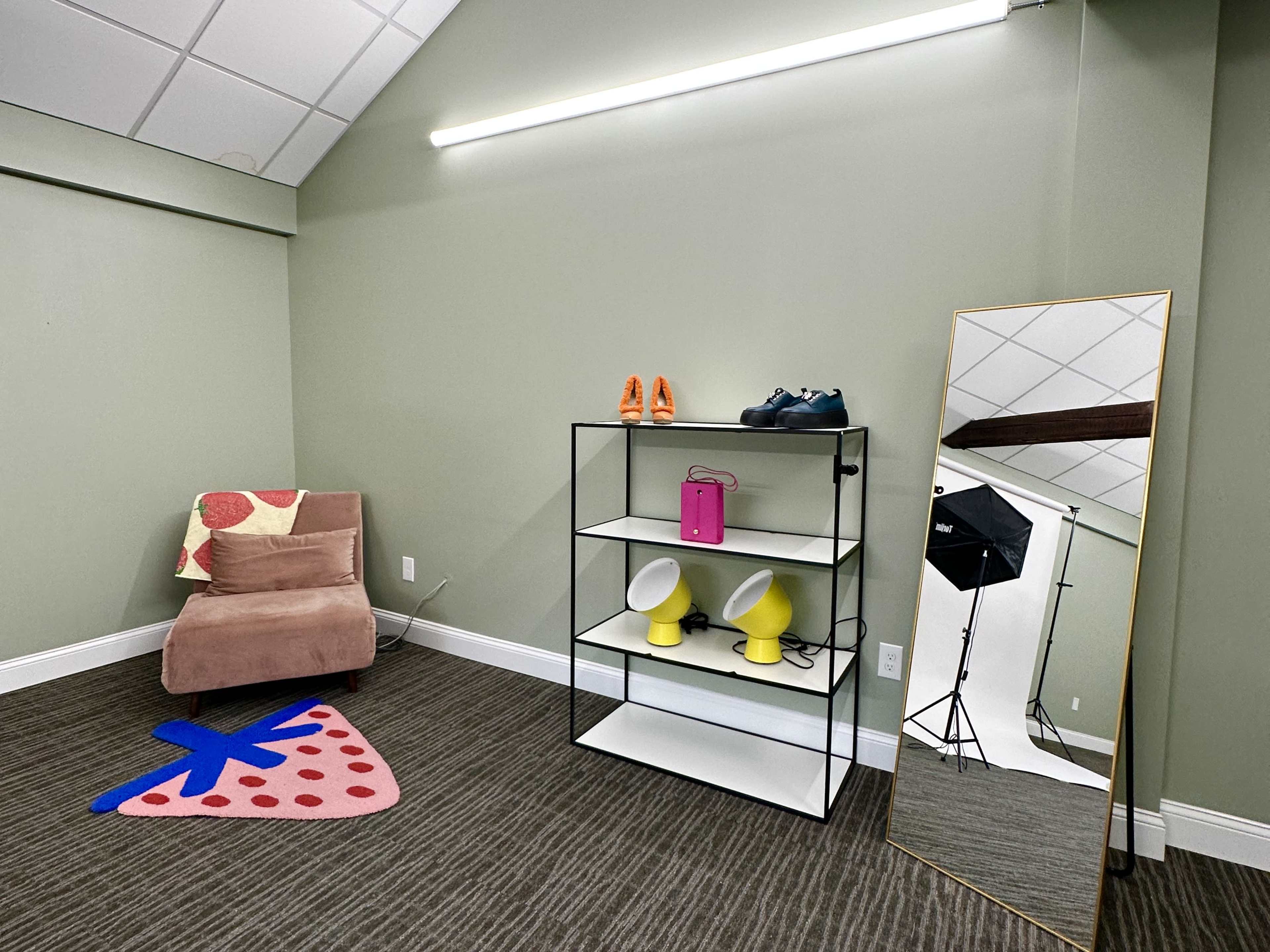 A room with a green wall, a brown chair with a fruit-patterned pillow, a black shelf displaying colorful shoes and accessories, and a large mirror beside a photography setup.