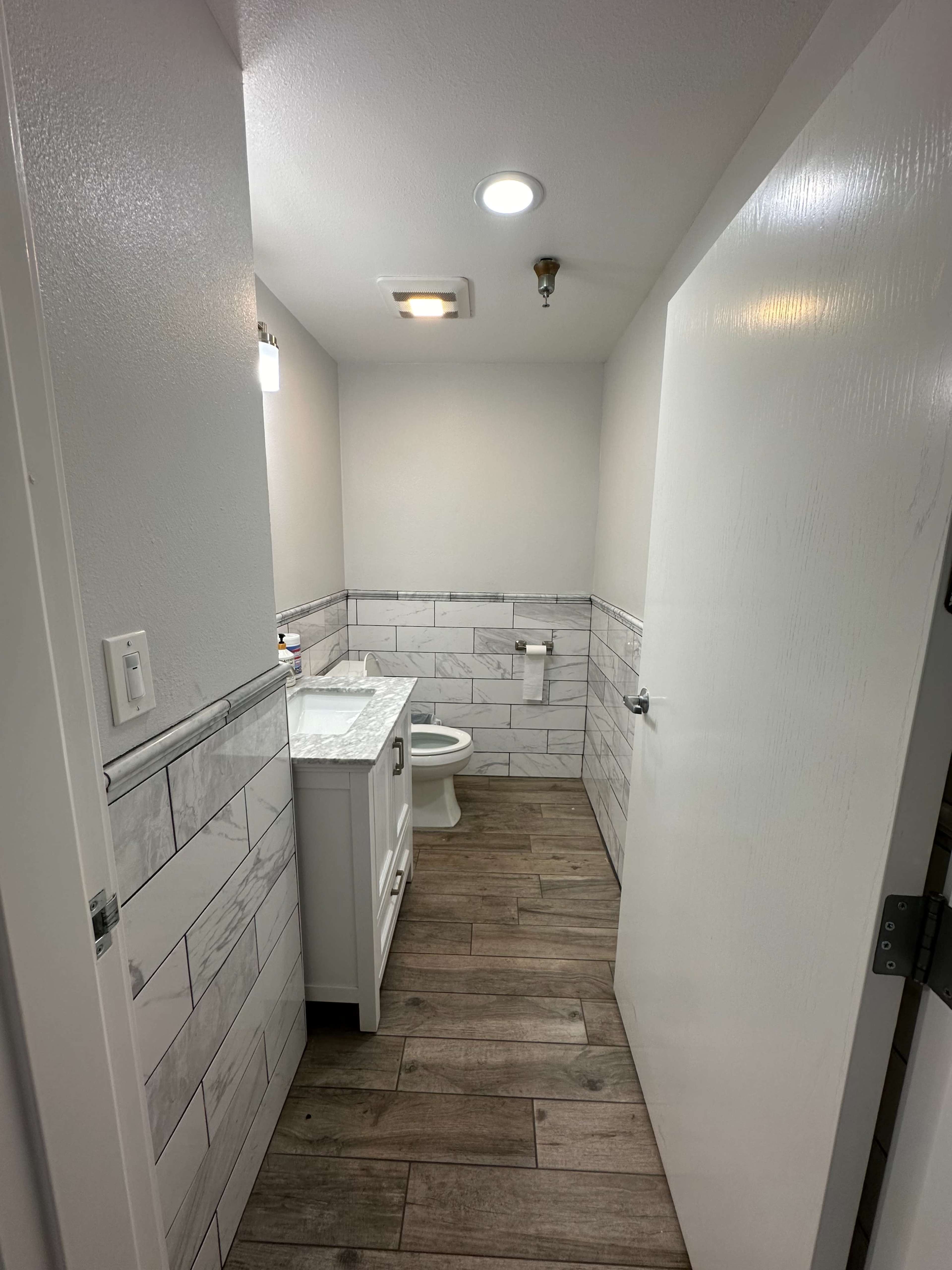 The image shows a modern bathroom with a white vanity, a toilet, and tiled walls, featuring wood-like flooring.