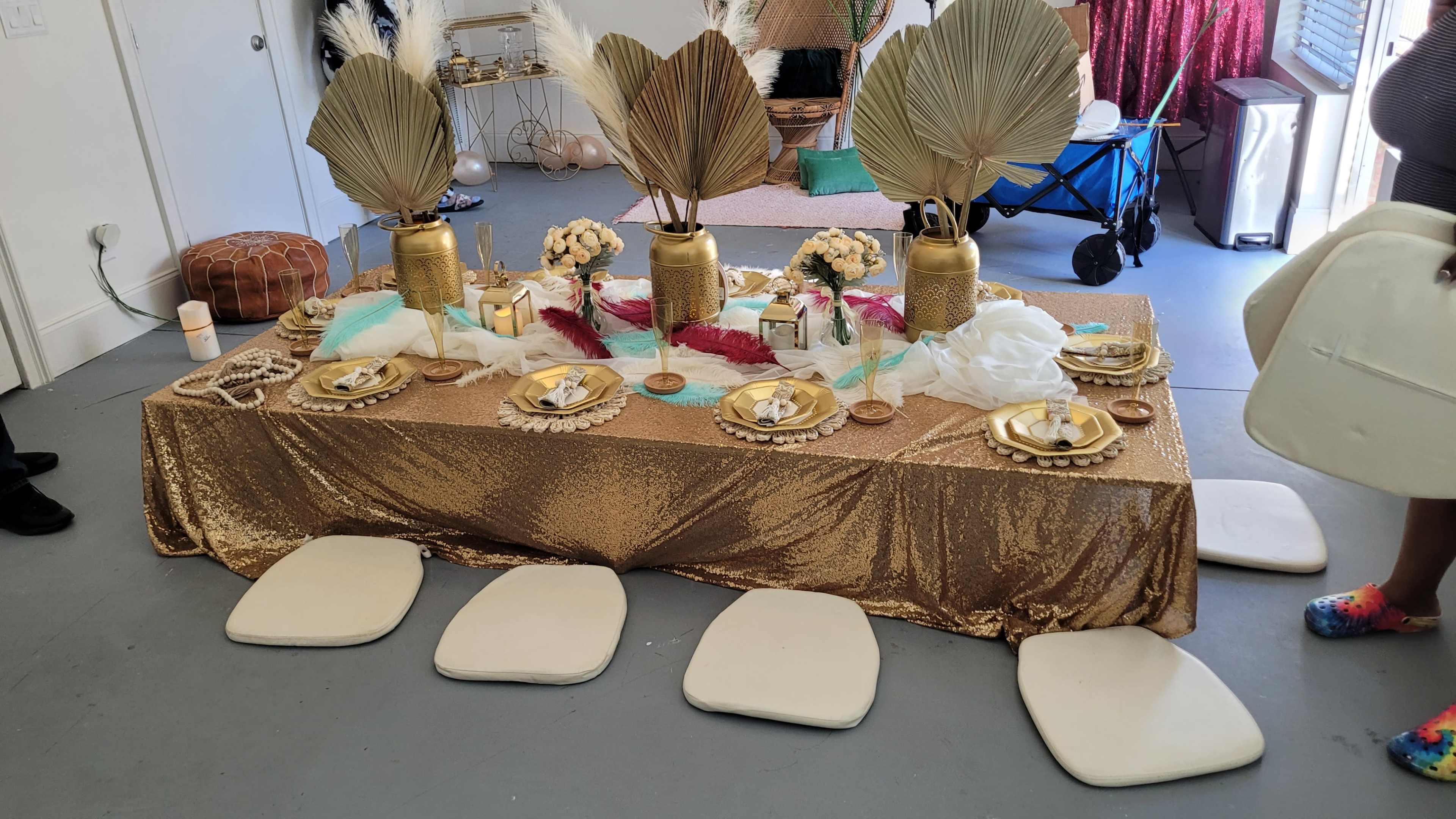 A gold tablecloth covers a decorated table set with plates, candles, and floral arrangements, surrounded by white seating cushions.