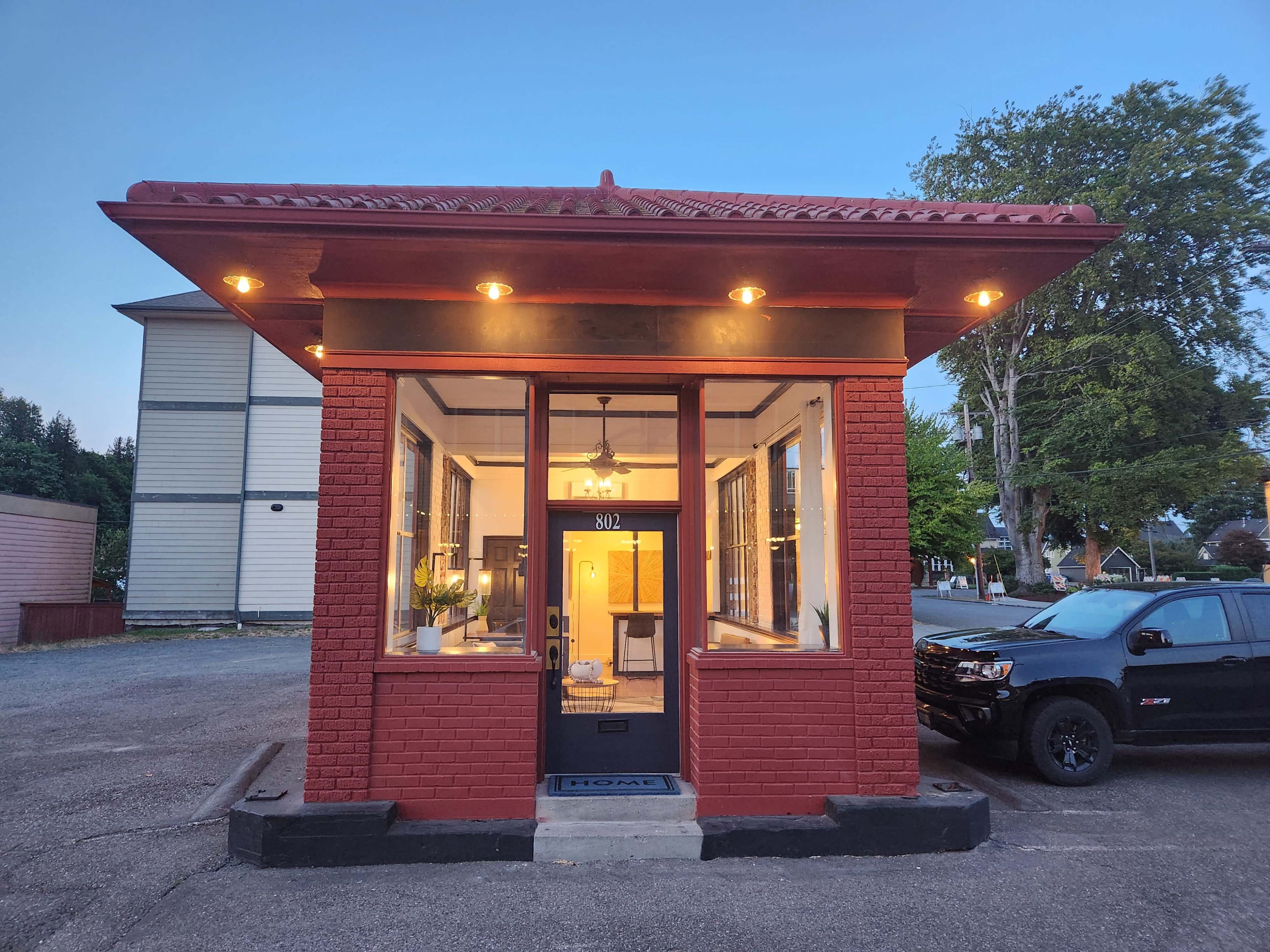 A red-brick building with a decorative roof, large windows, and a welcoming front entrance, situated in a parking area with a black vehicle nearby.