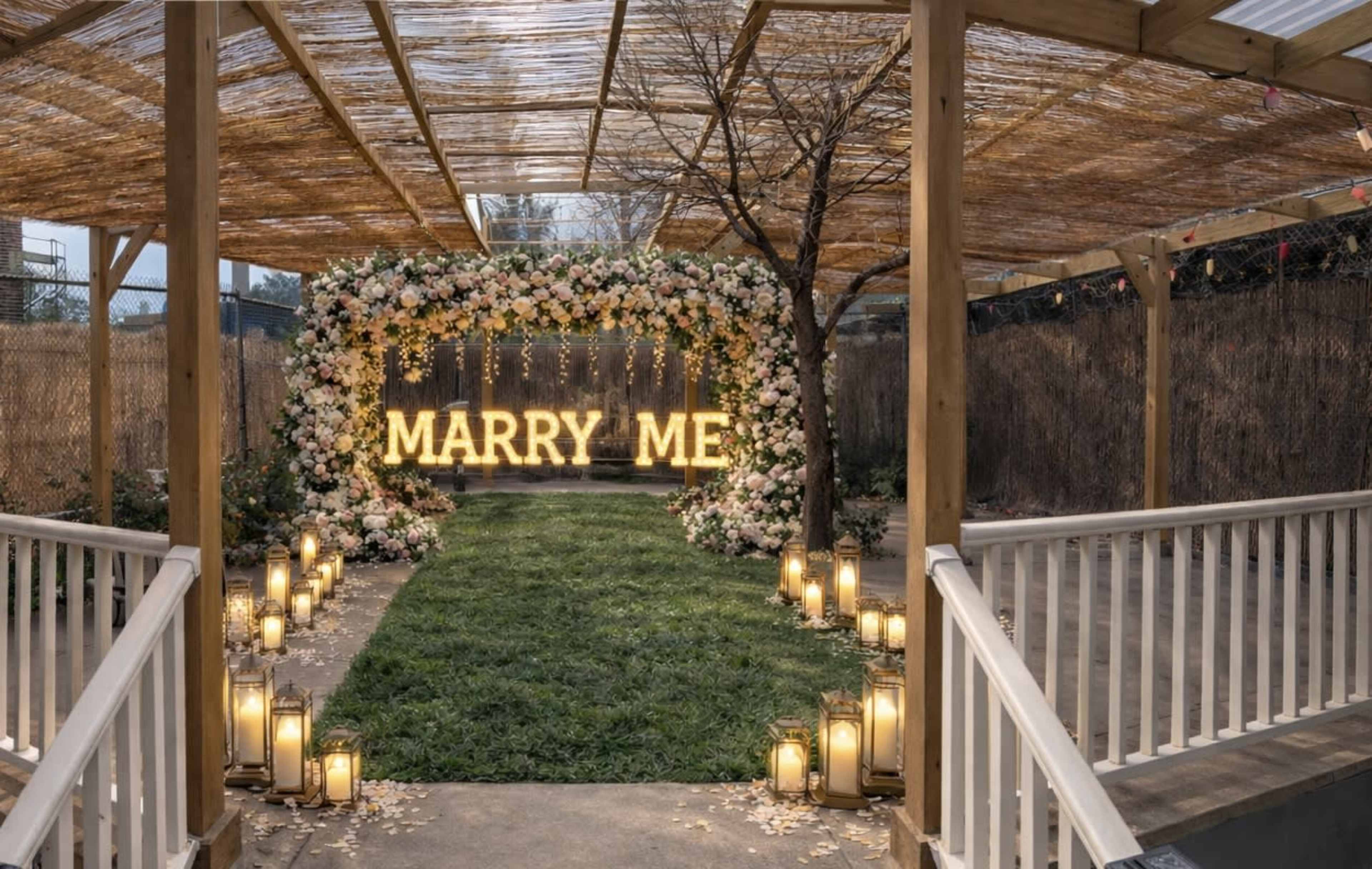 A wedding proposal setup features a floral arch with large illuminated letters spelling "MARRY ME," surrounded by lanterns along a grassy path.