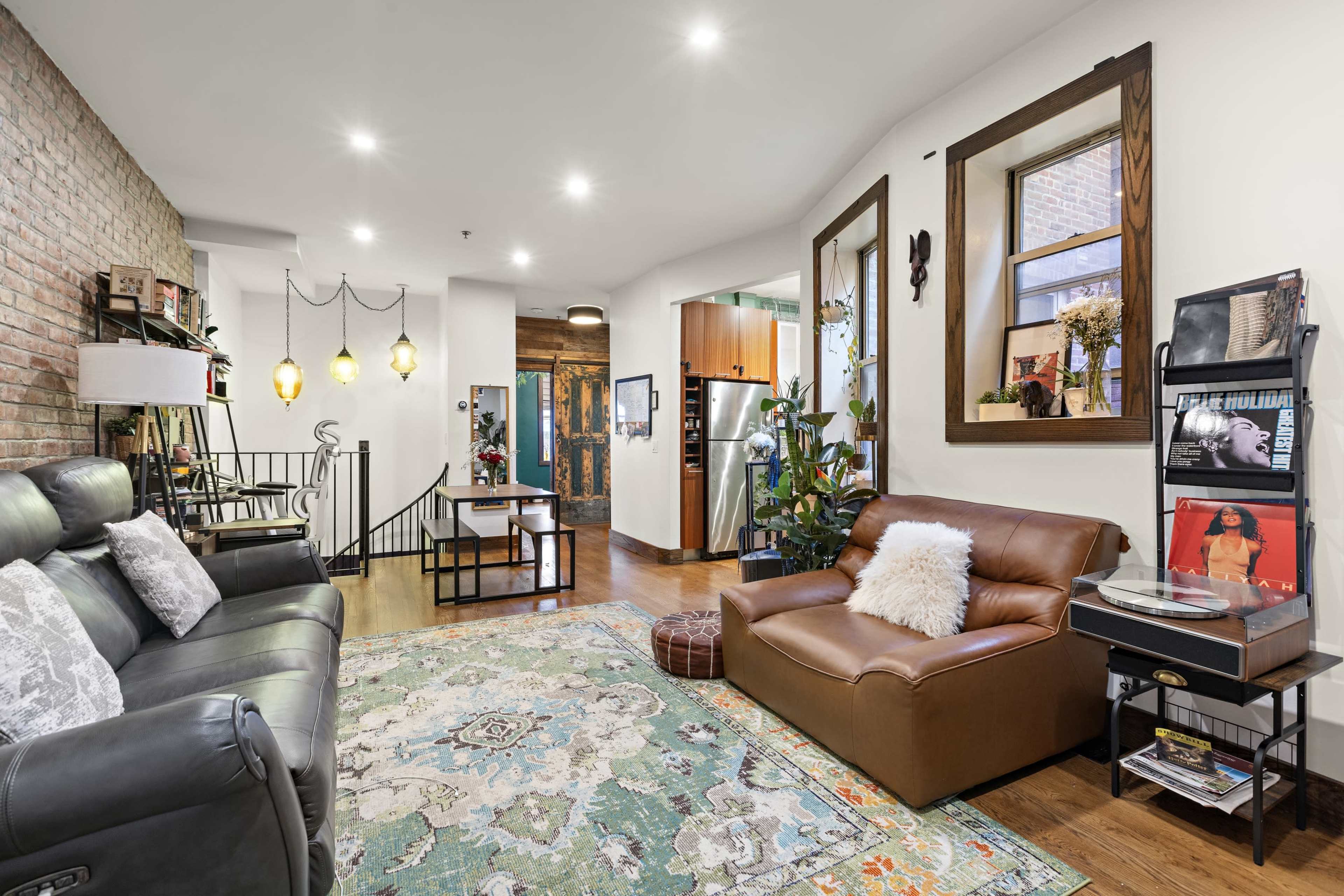 The image shows a cozy living room with brick walls, large windows, leather seating, and a decorative area rug.