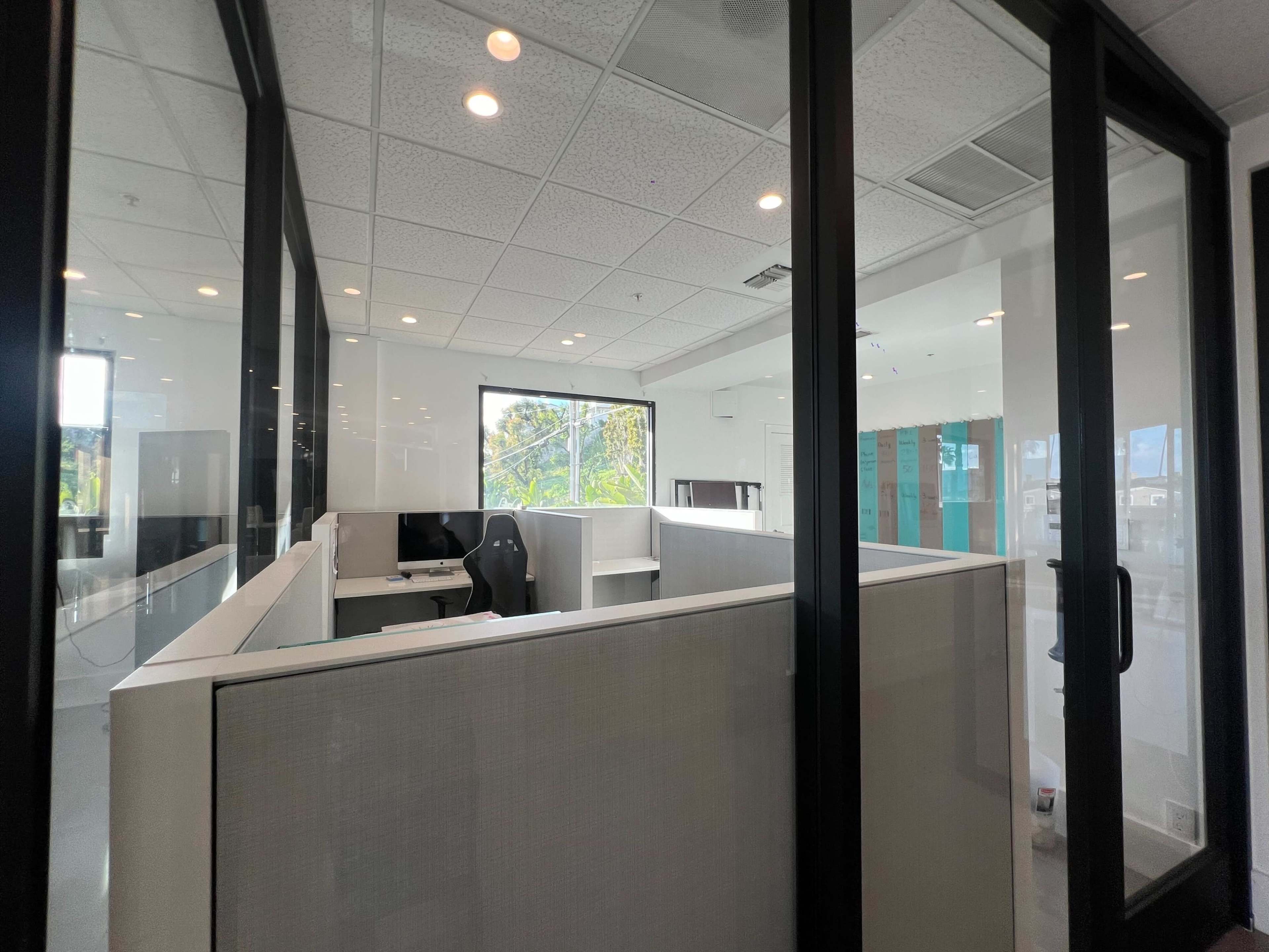A modern office space with glass partitions, a desk with a computer, and a large window overlooking greenery.