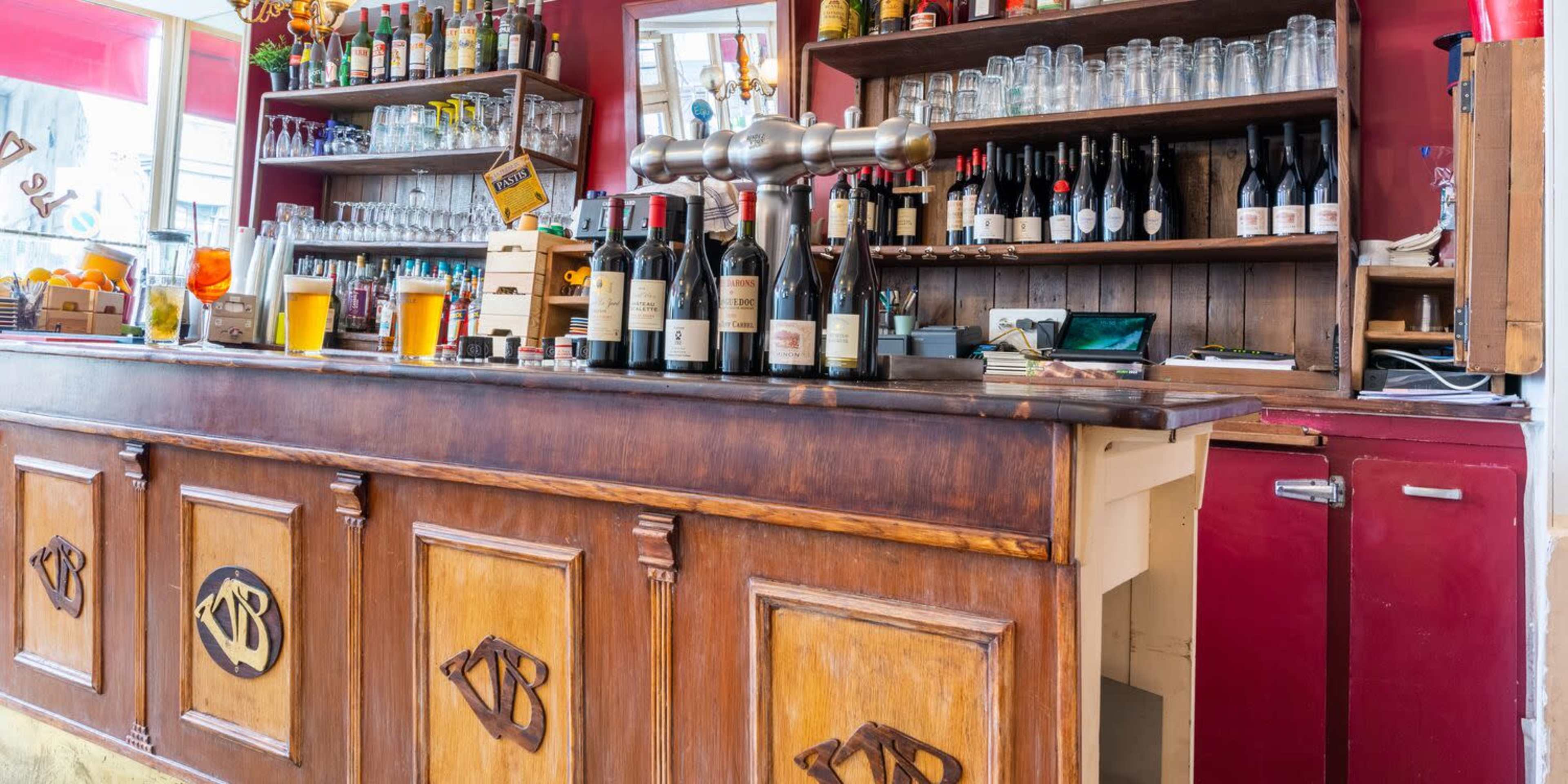 The image shows a wooden bar with several bottles of wine, glassware, and a row of beer taps behind it, set in a brightly lit room with red walls.