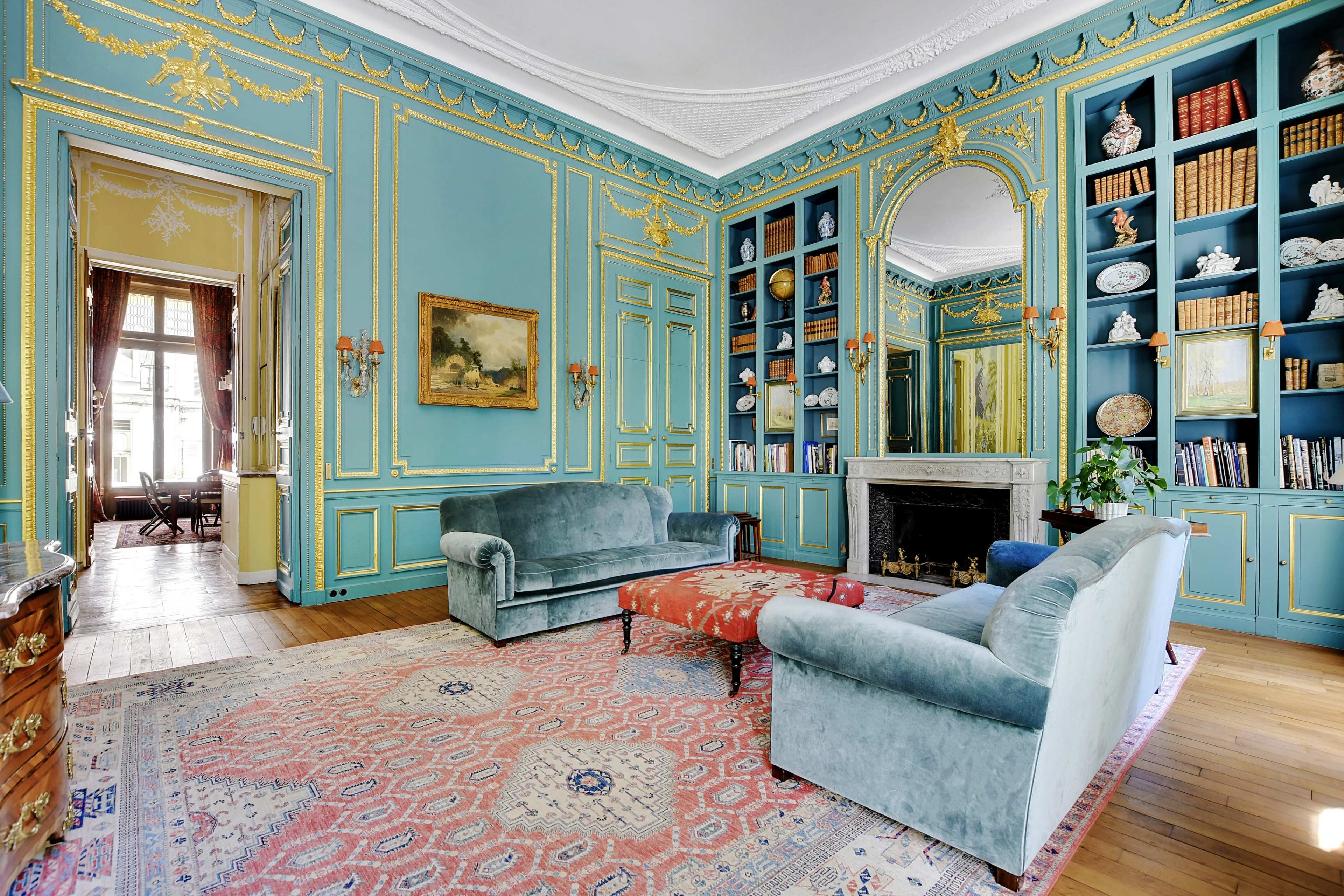A spacious living room features light blue walls with ornate golden detailing, two green sofas, and a decorative bookshelf filled with books and artifacts.