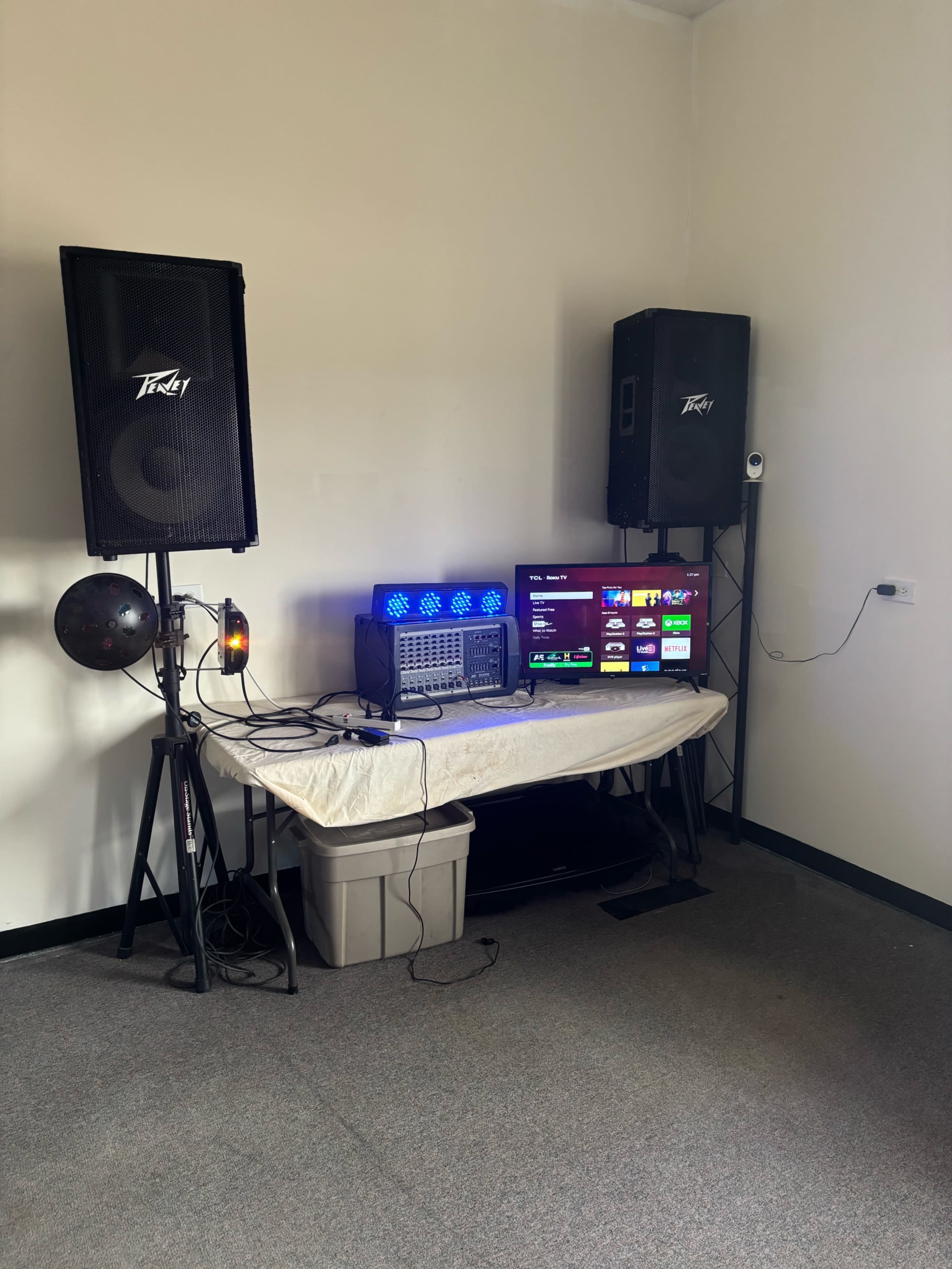 The image shows a music setup featuring two large speakers, a sound mixer, a television displaying a menu, and various audio equipment arranged on a table against a plain wall.