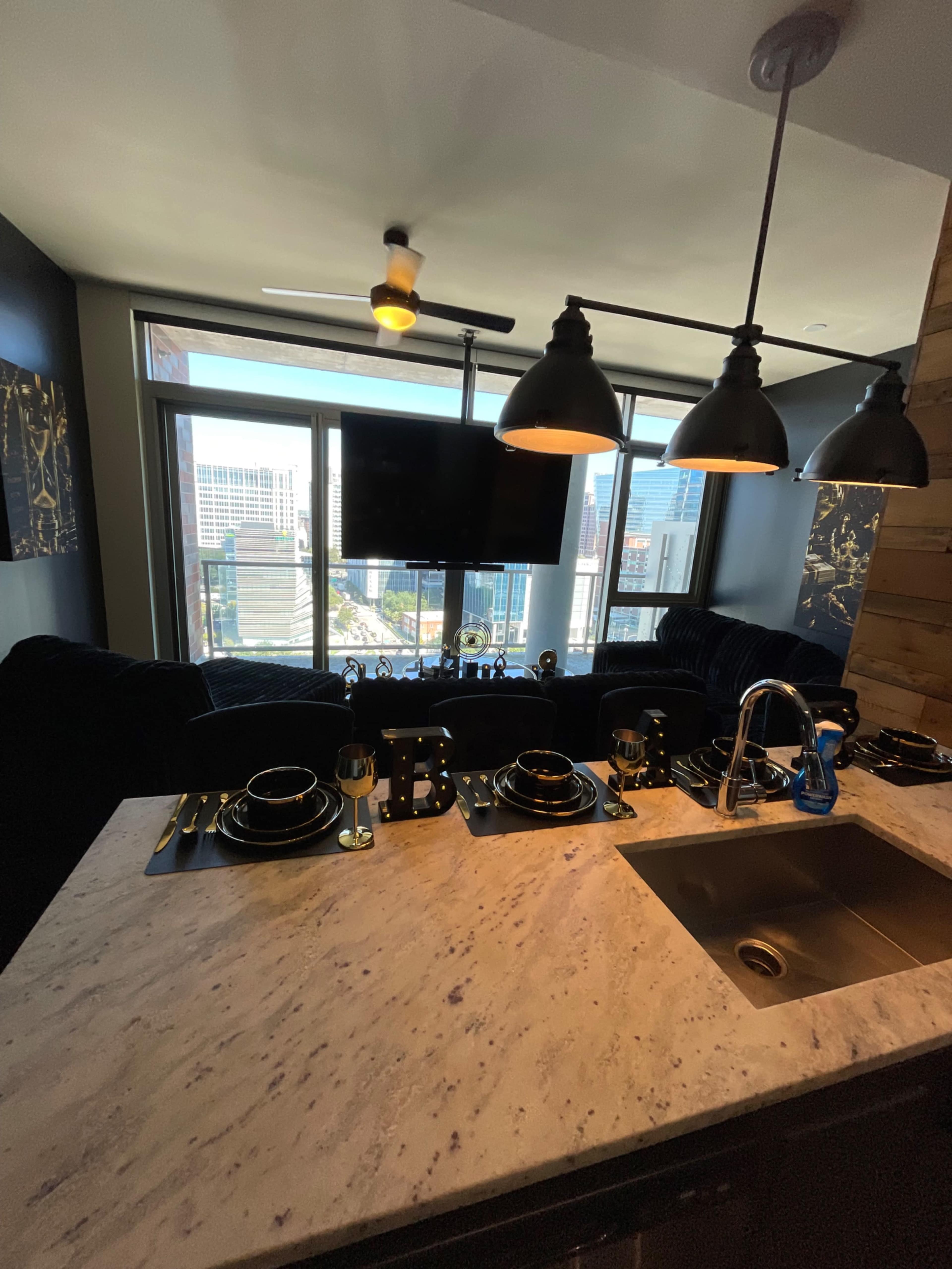 A modern kitchen with a marble countertop features a dining setup for four, with a large window showcasing a city view in the background.