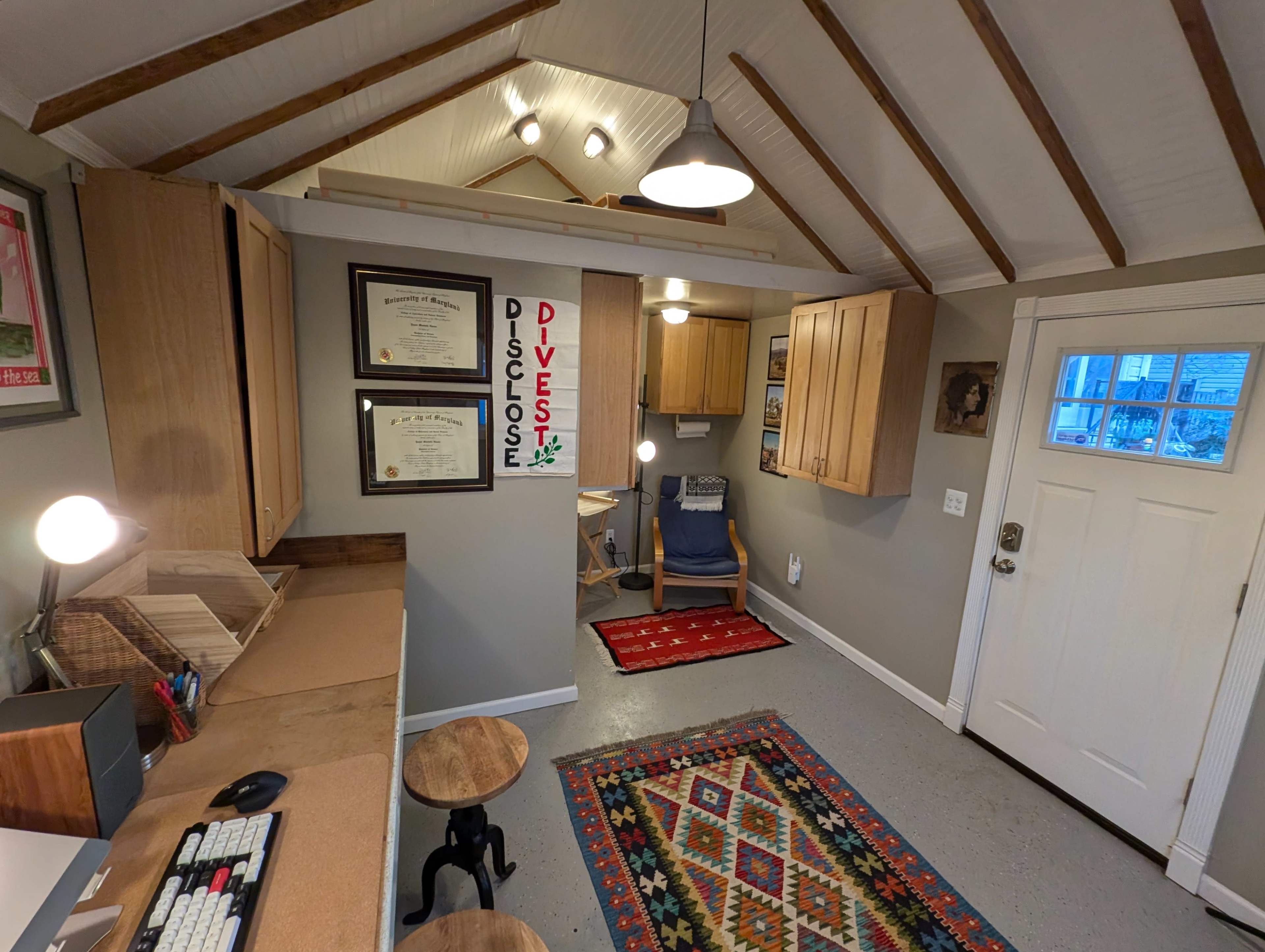 The image shows a small office space with wooden beams on the ceiling, a desk with a computer, shelves with framed certificates, and a door leading outside.