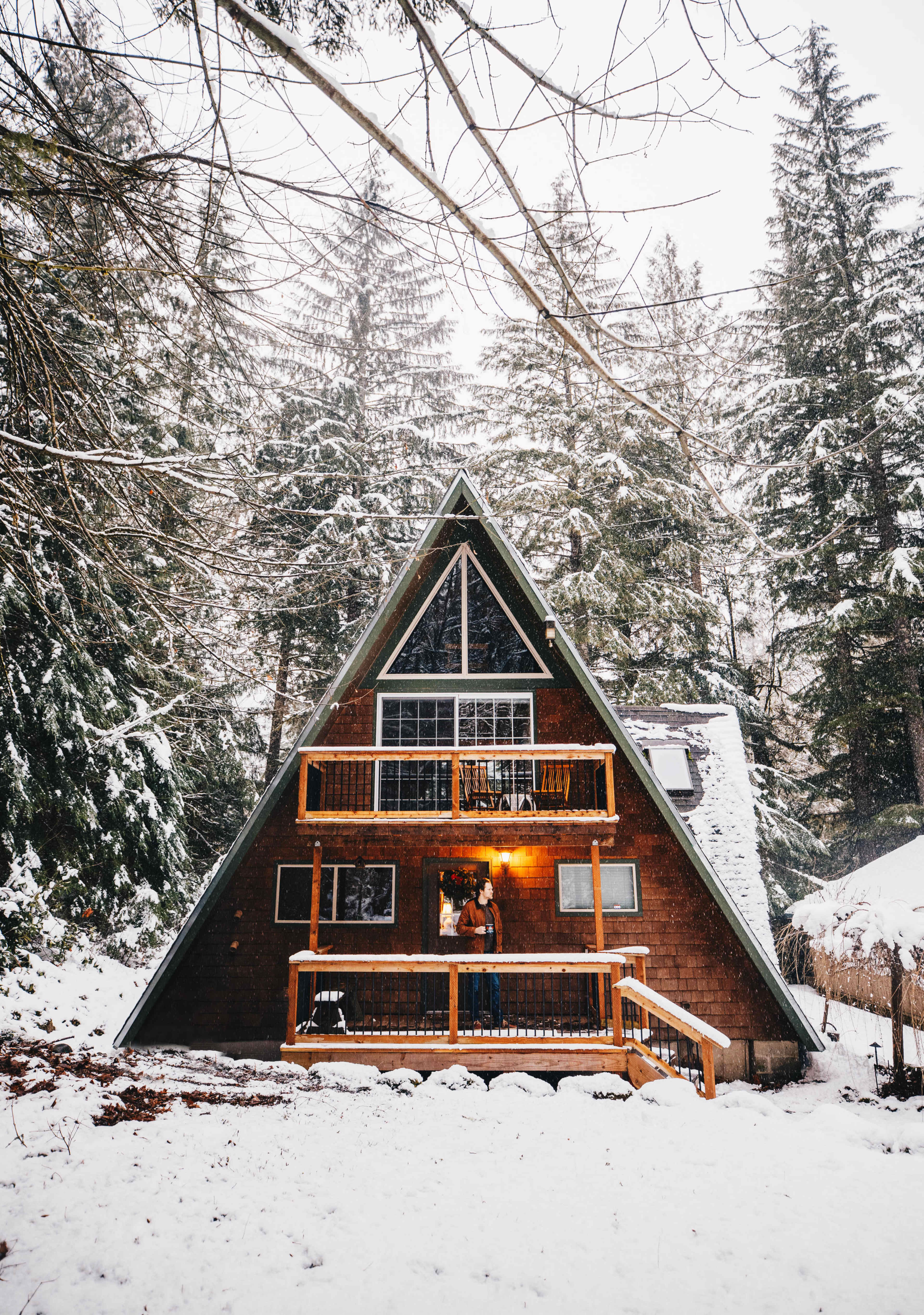 A wooden A-frame cabin with a large porch is surrounded by snow-covered trees in a winter landscape.