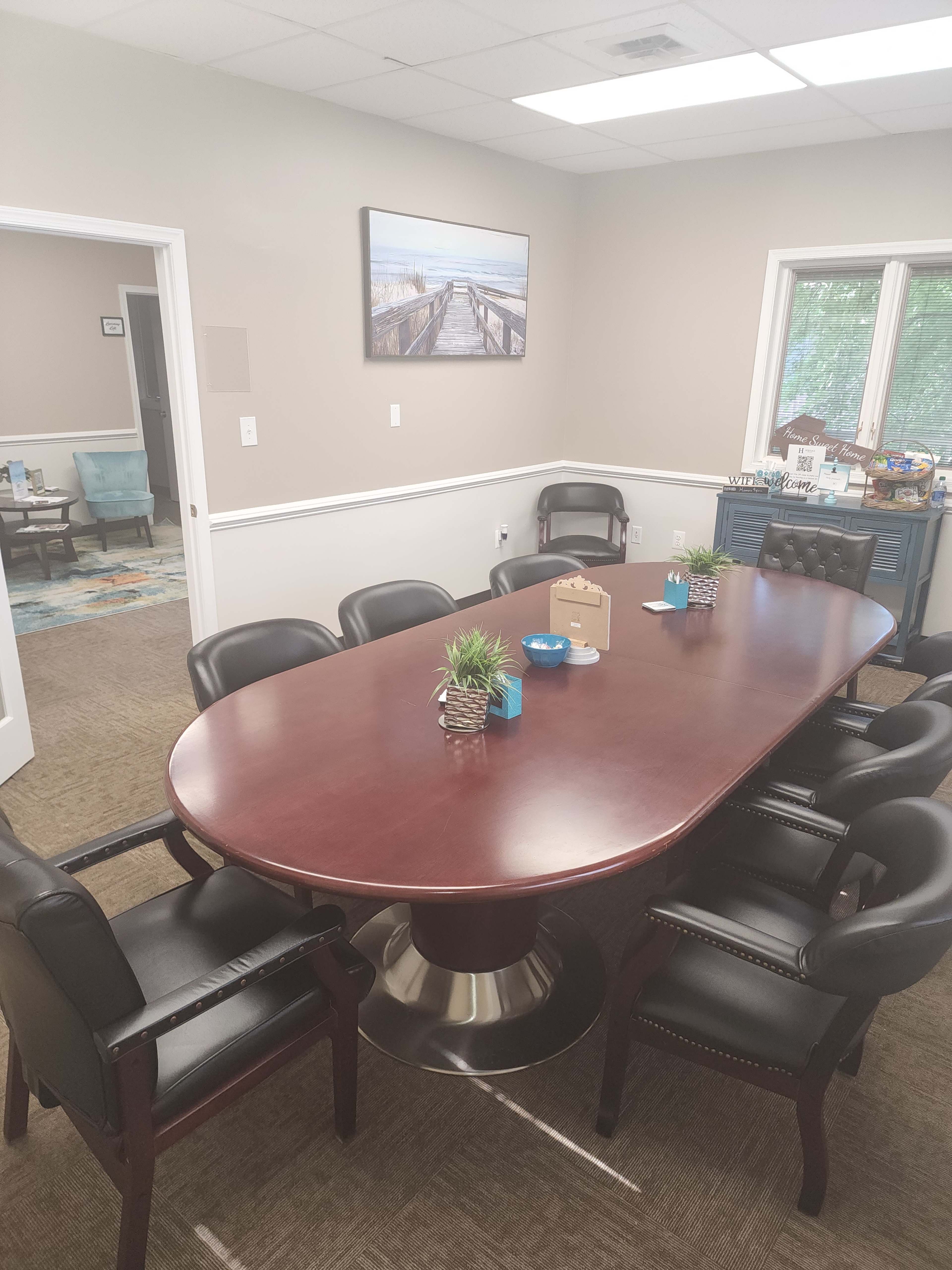 A conference room with a large oval table surrounded by black leather chairs, a painting on the wall, and a side area with refreshments.