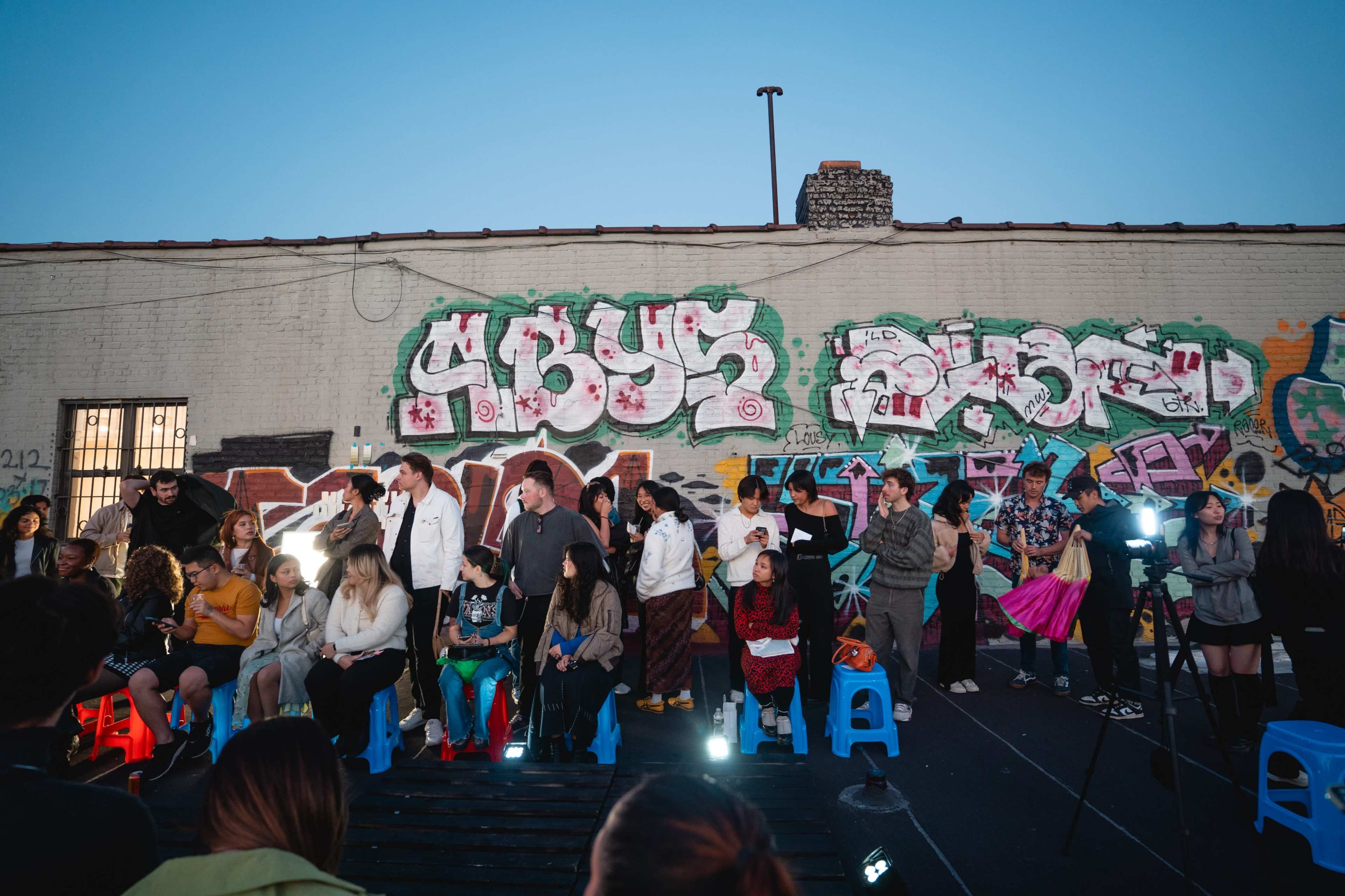 A diverse group of people sits on blue chairs in front of a graffiti-covered wall during an outdoor event.
