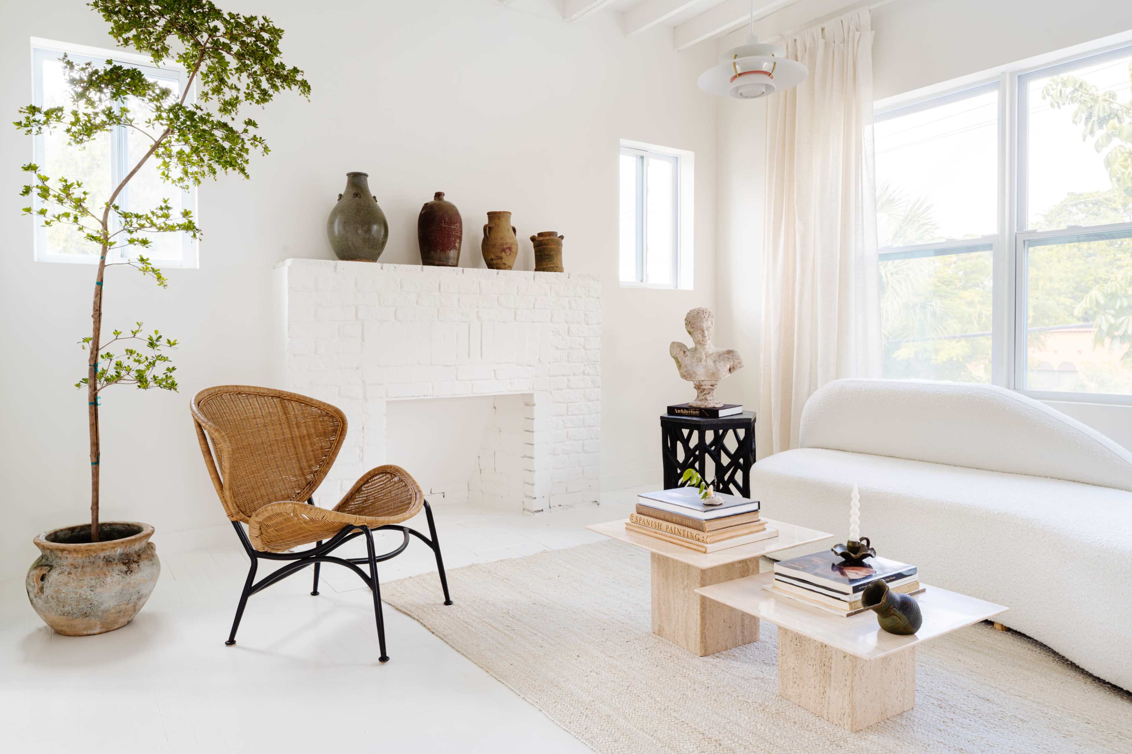 A minimalist living room features a white sofa, a rattan chair, ceramic vases, and two wooden side tables with stacked books.