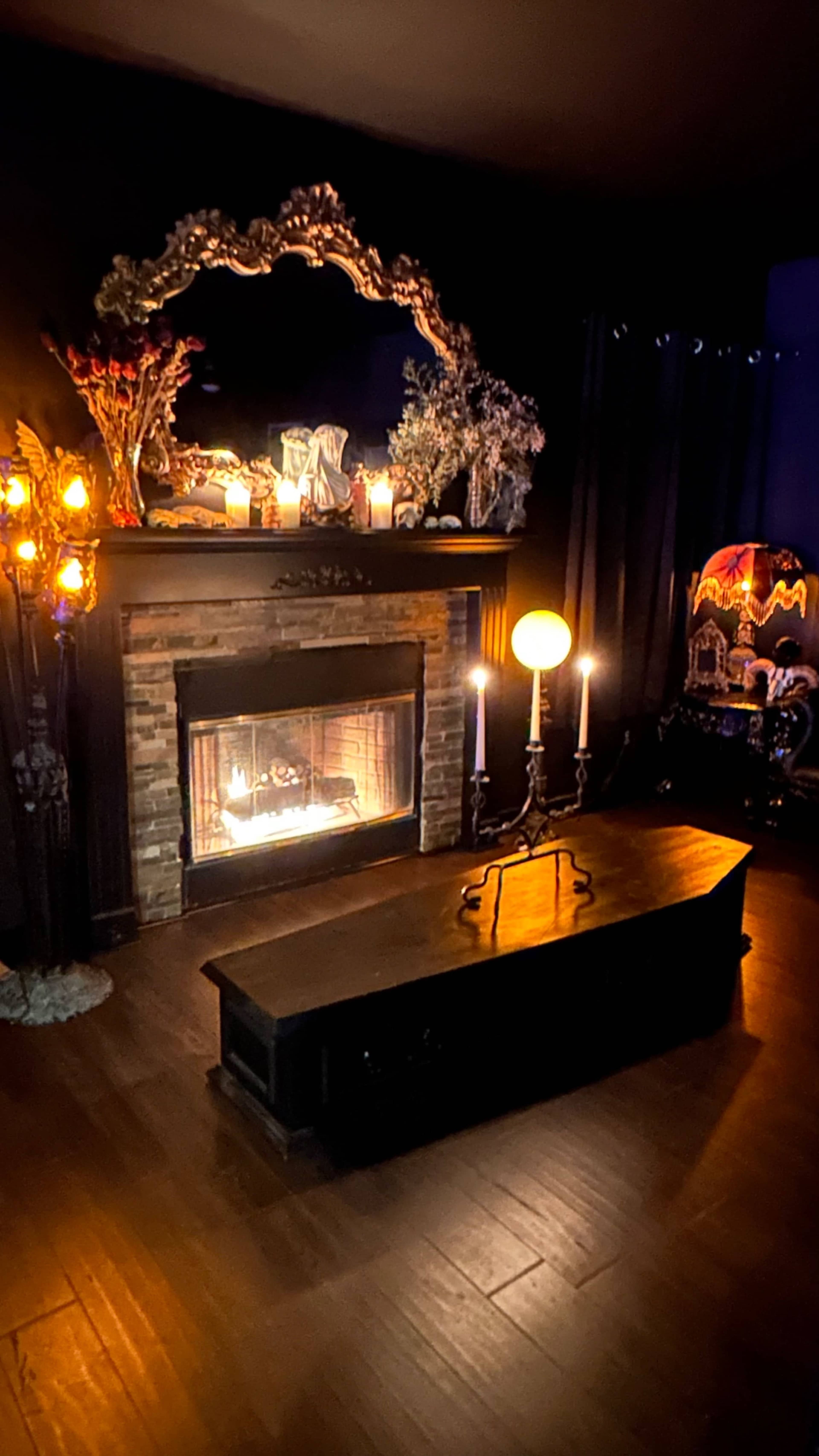 A dimly lit room featuring a fireplace with a flickering flame, a large ornate mirror above it, candles arranged on the mantel, and a wooden coffee table in front.