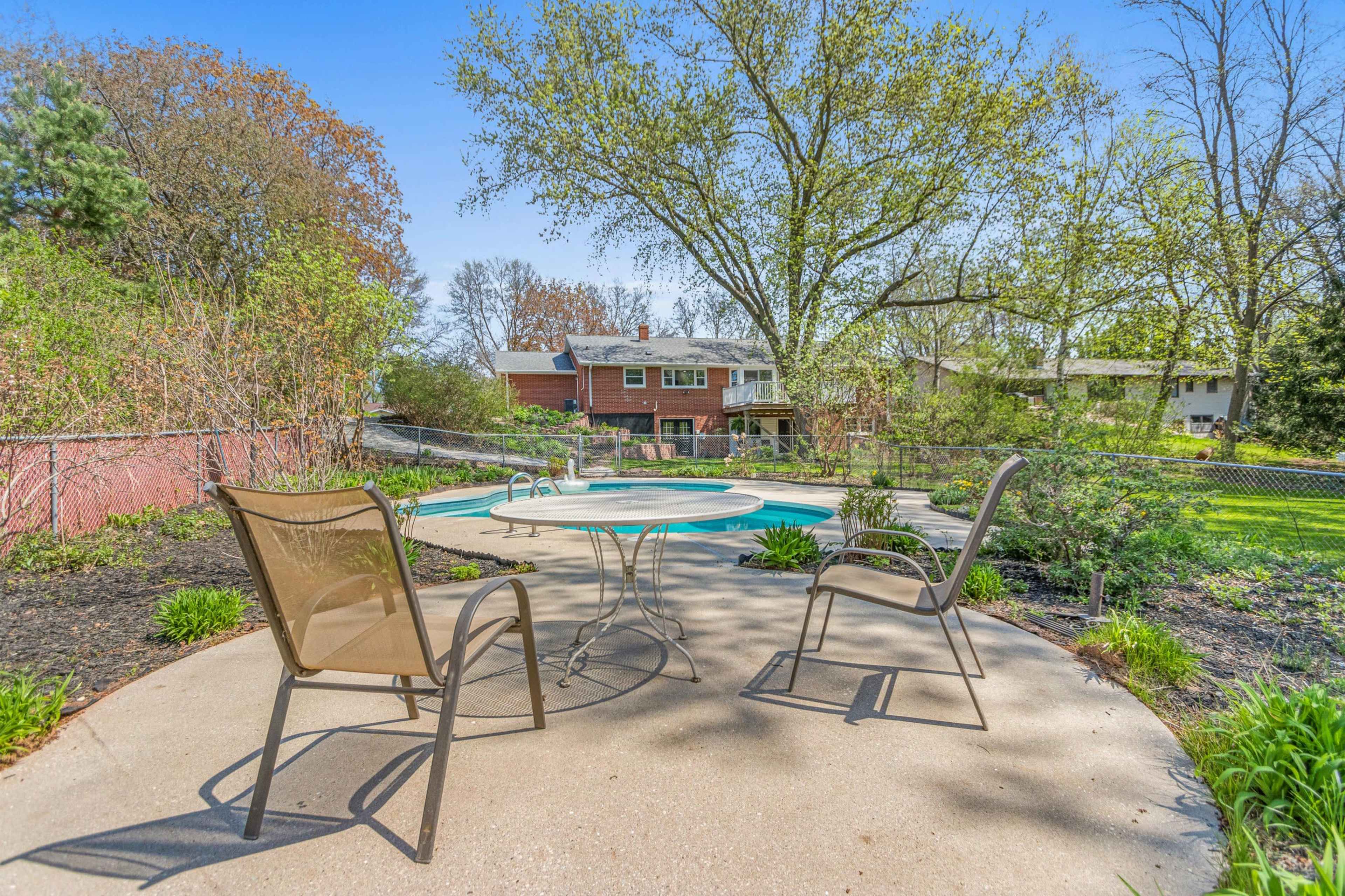A concrete patio with two chairs overlooks a swimming pool surrounded by greenery and residential buildings in the background.