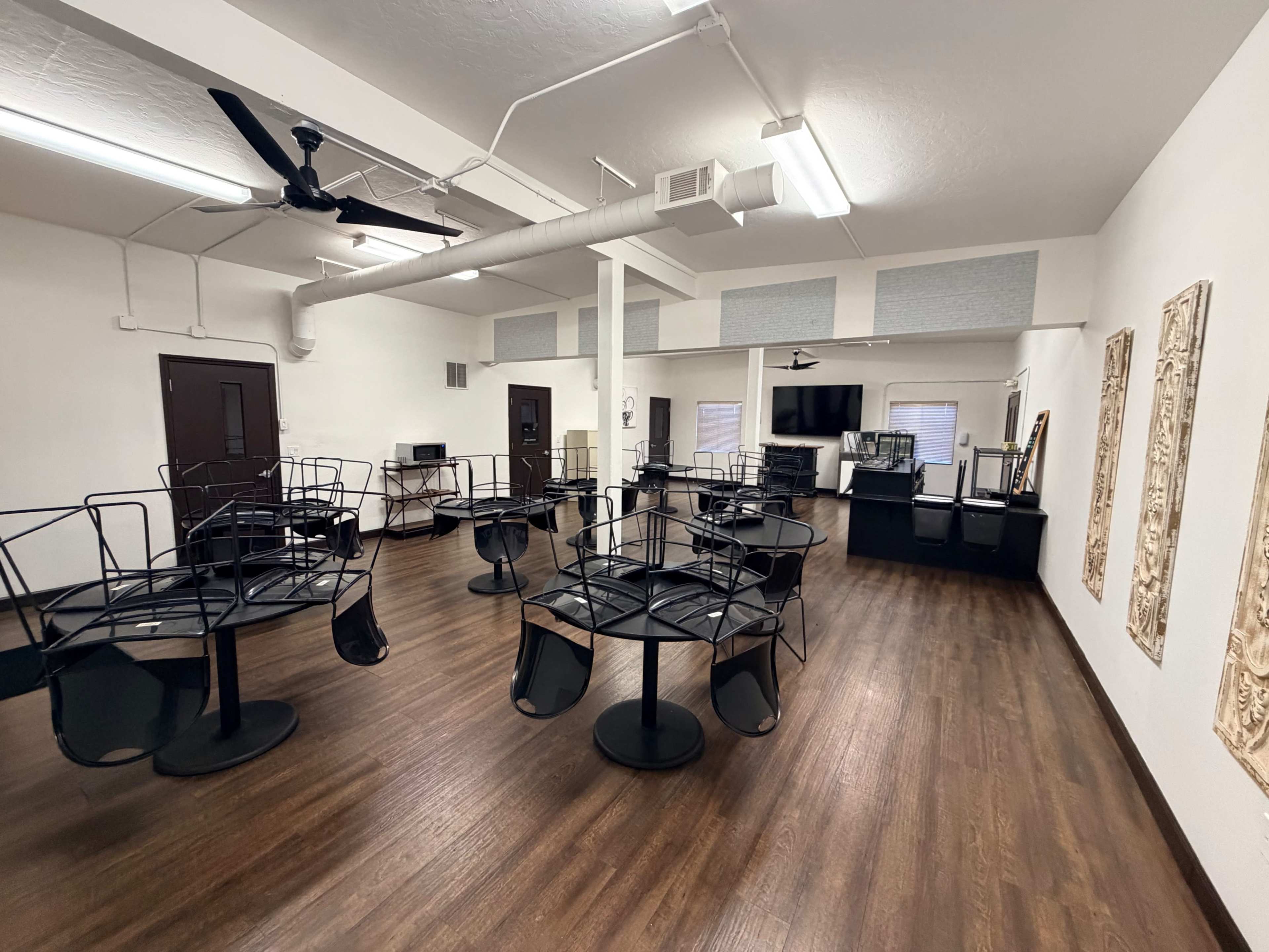 The image shows an empty room with several black tables and chairs stacked on their sides, wooden flooring, and a small kitchen area against one wall.