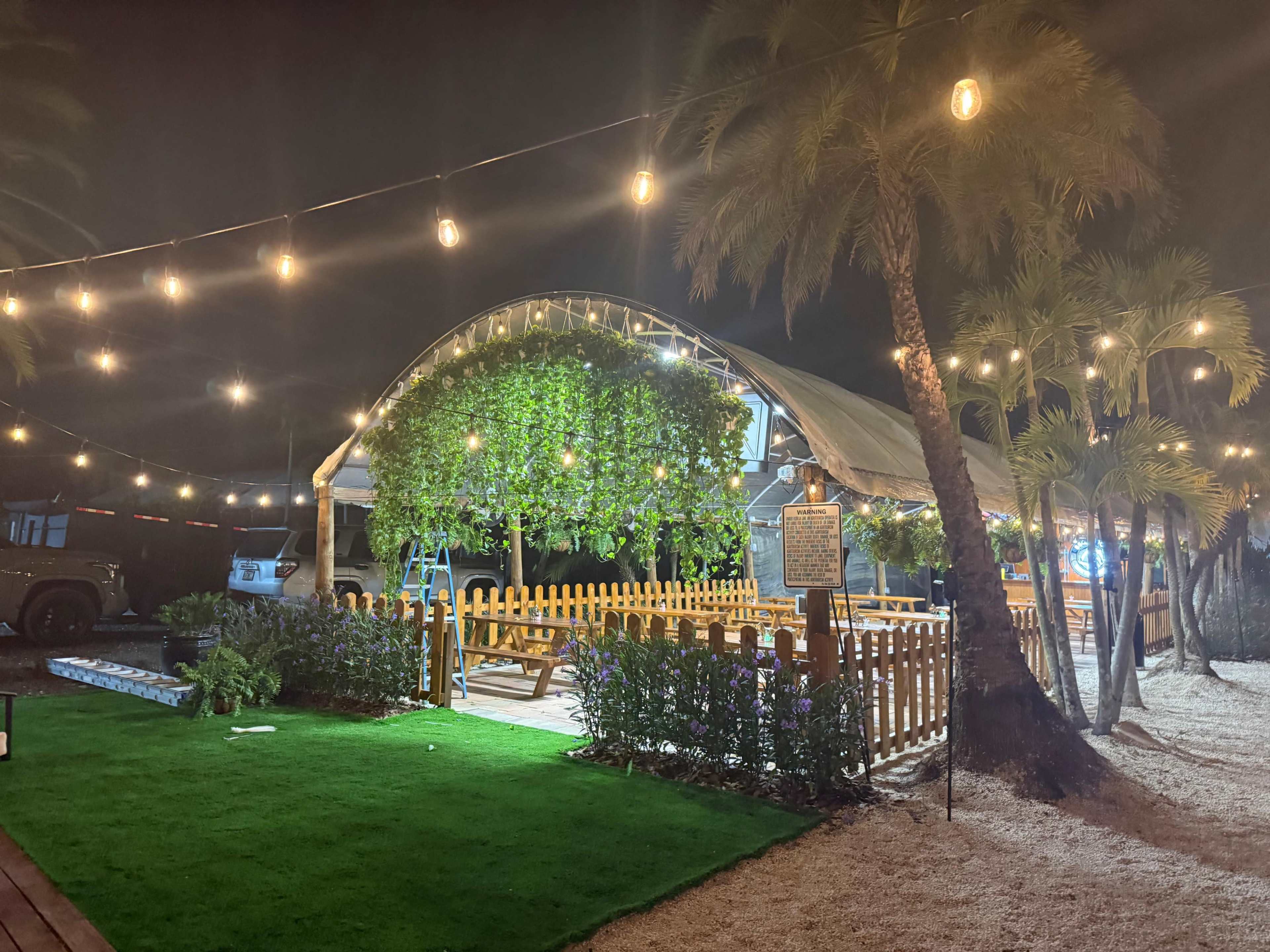 A canopy-covered outdoor seating area is illuminated by string lights and surrounded by tropical plants.