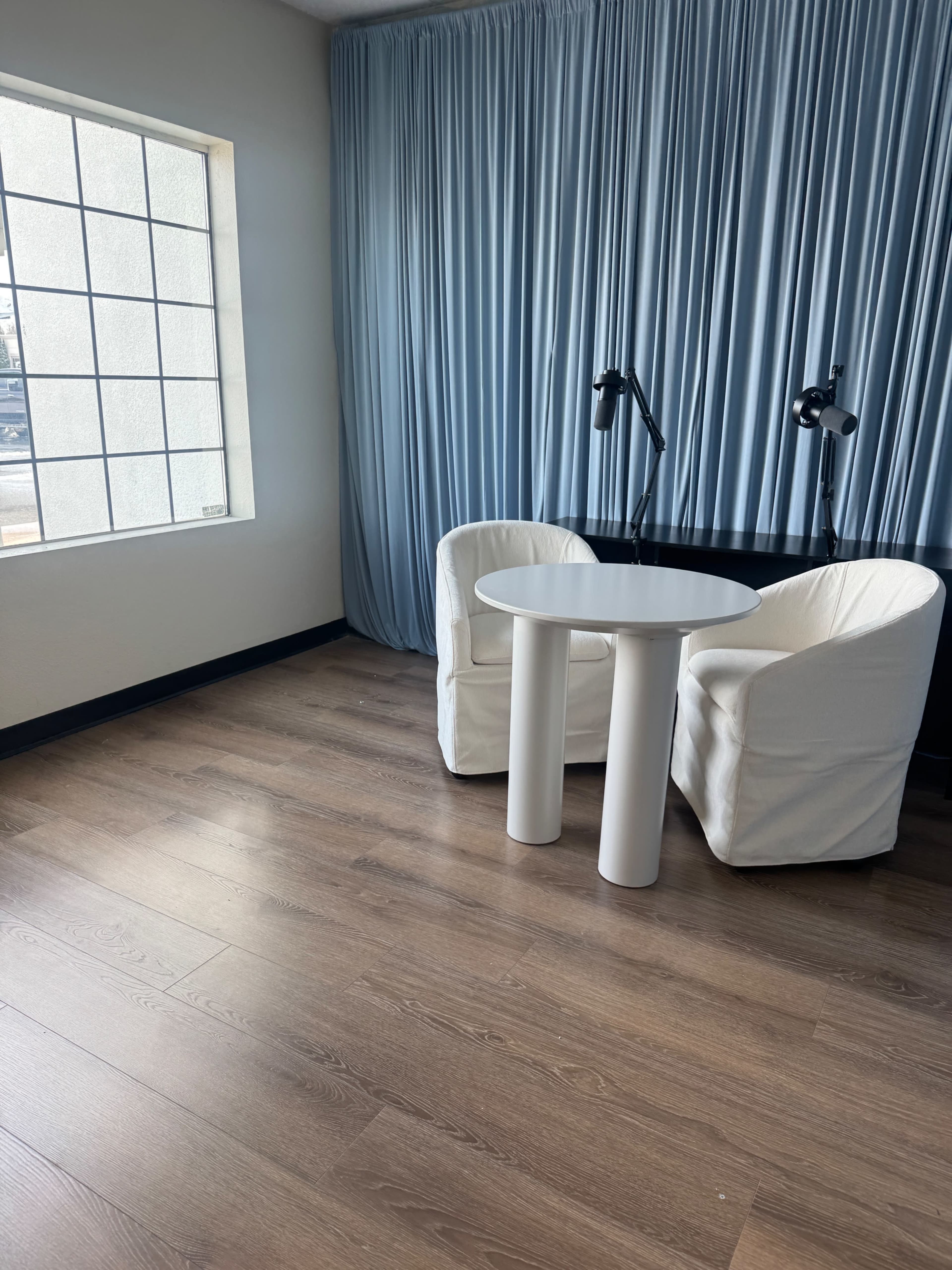 The image shows a minimalistic room featuring two white armchairs and a round white table, with a large window and light blue curtains in the background.