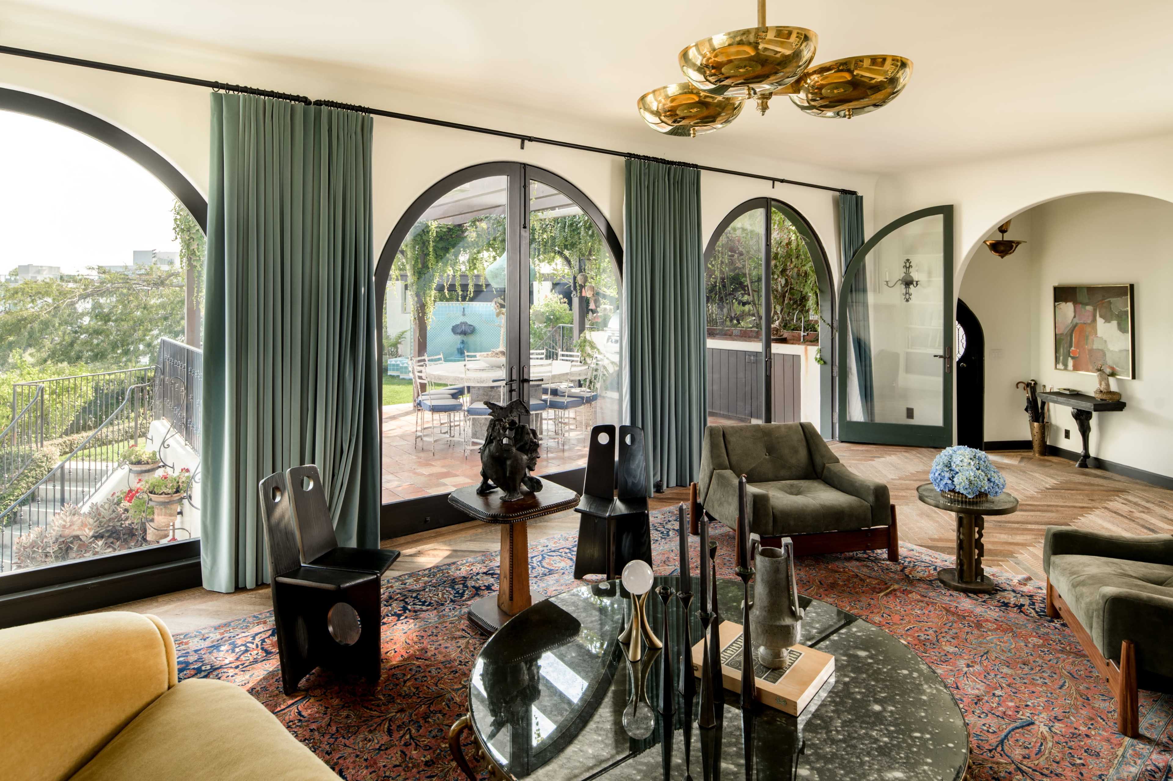 The image shows a modern living room with large arched windows overlooking a patio and pool, furnished with a round table and contemporary seating.