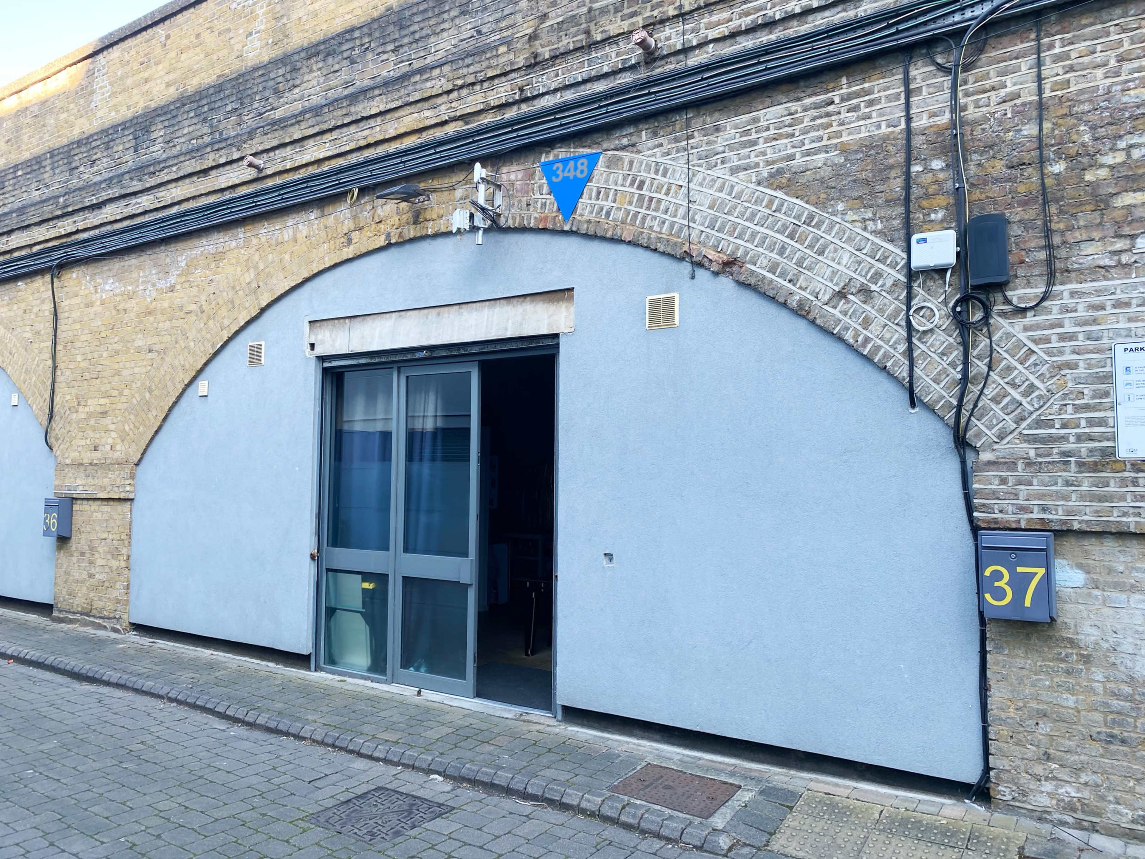 The image shows a gray building with an arched entrance, featuring a blue sign displaying the number 348 and adjacent mailbox numbers 37 on a cobblestone street.