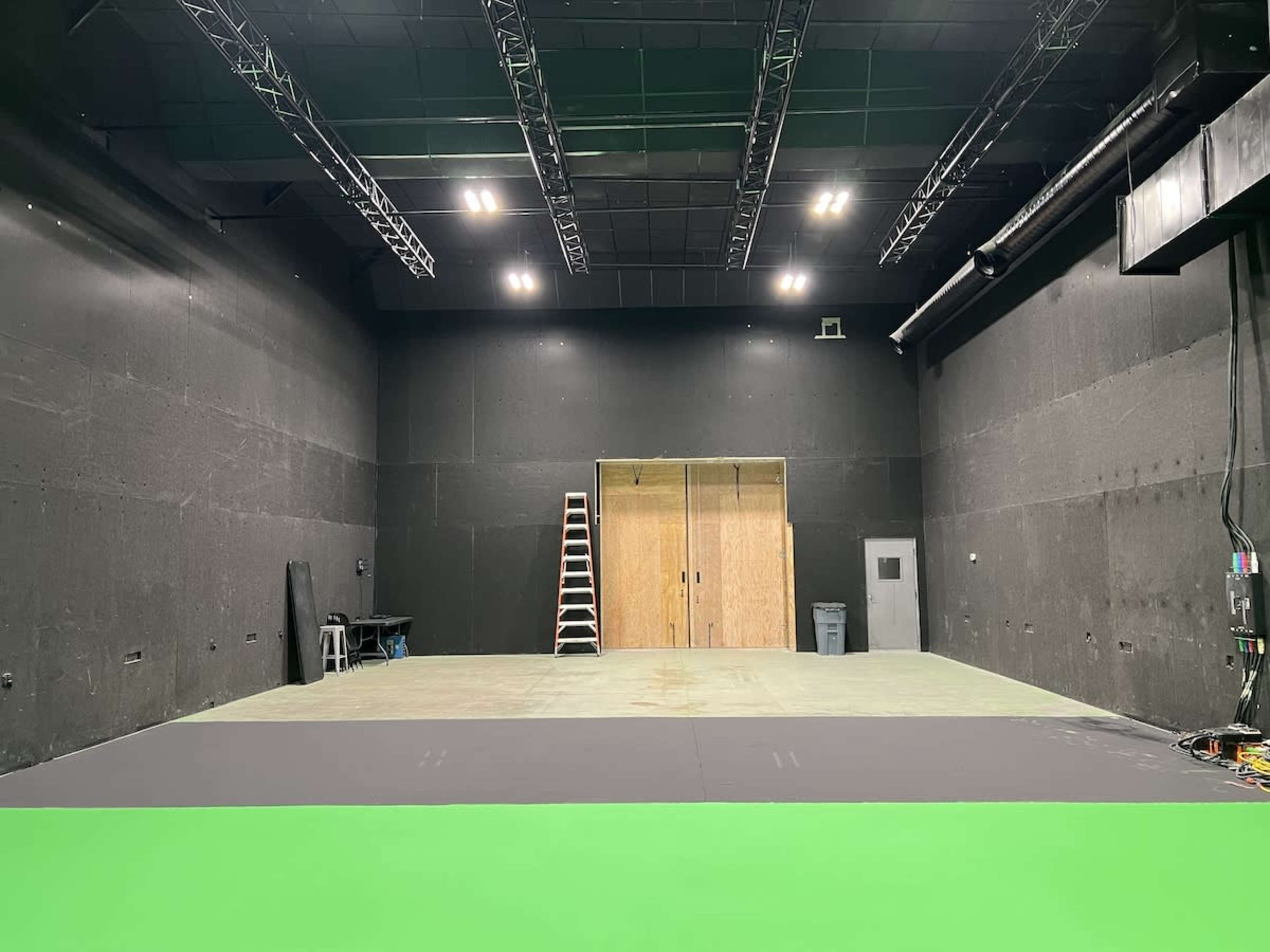 A large, empty studio space with dark walls, a green screen area in the foreground, and wooden doors at the back.