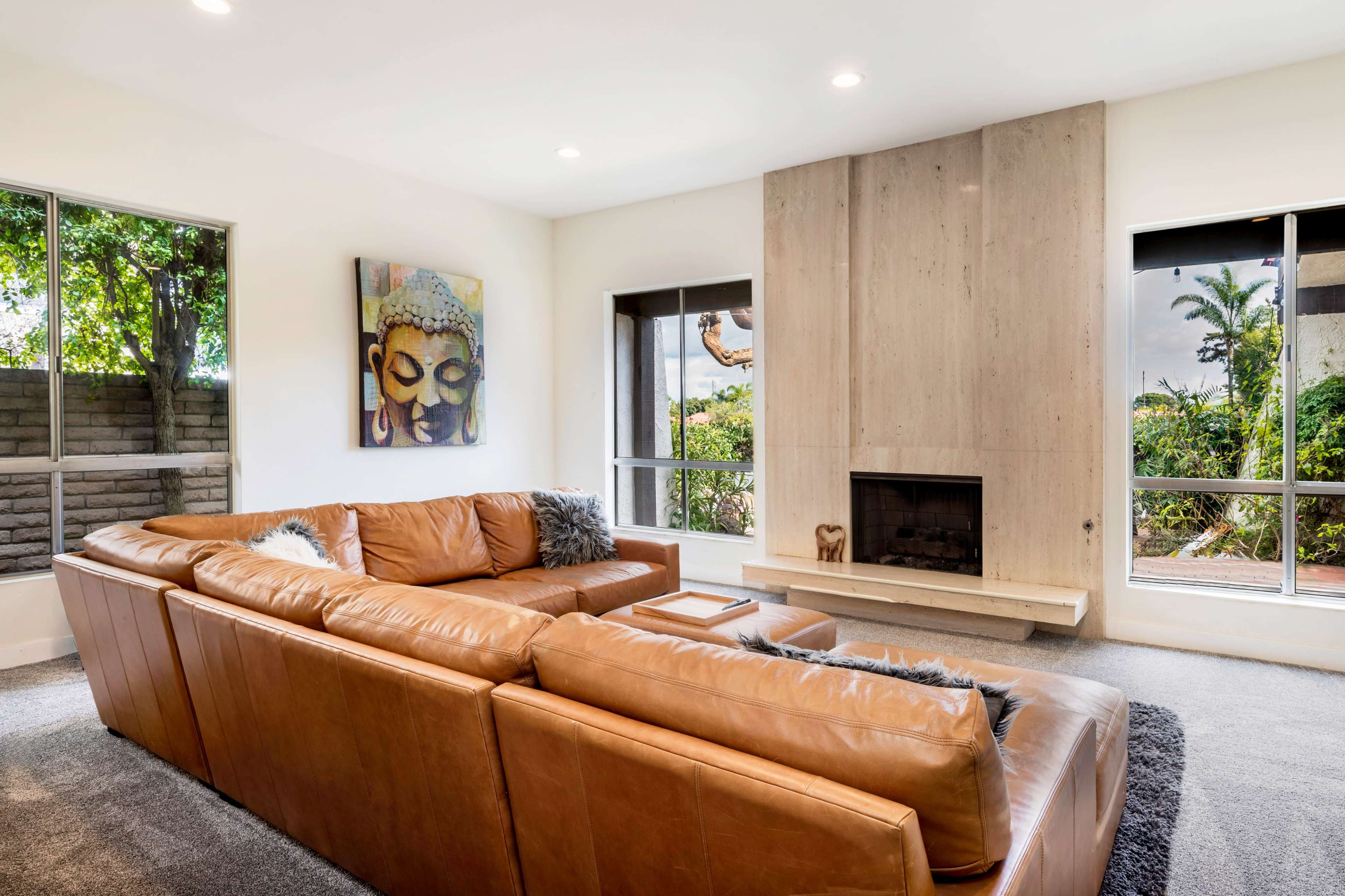 A spacious living room features a large brown leather sectional sofa, a minimalist stone fireplace, and large windows with views of greenery.