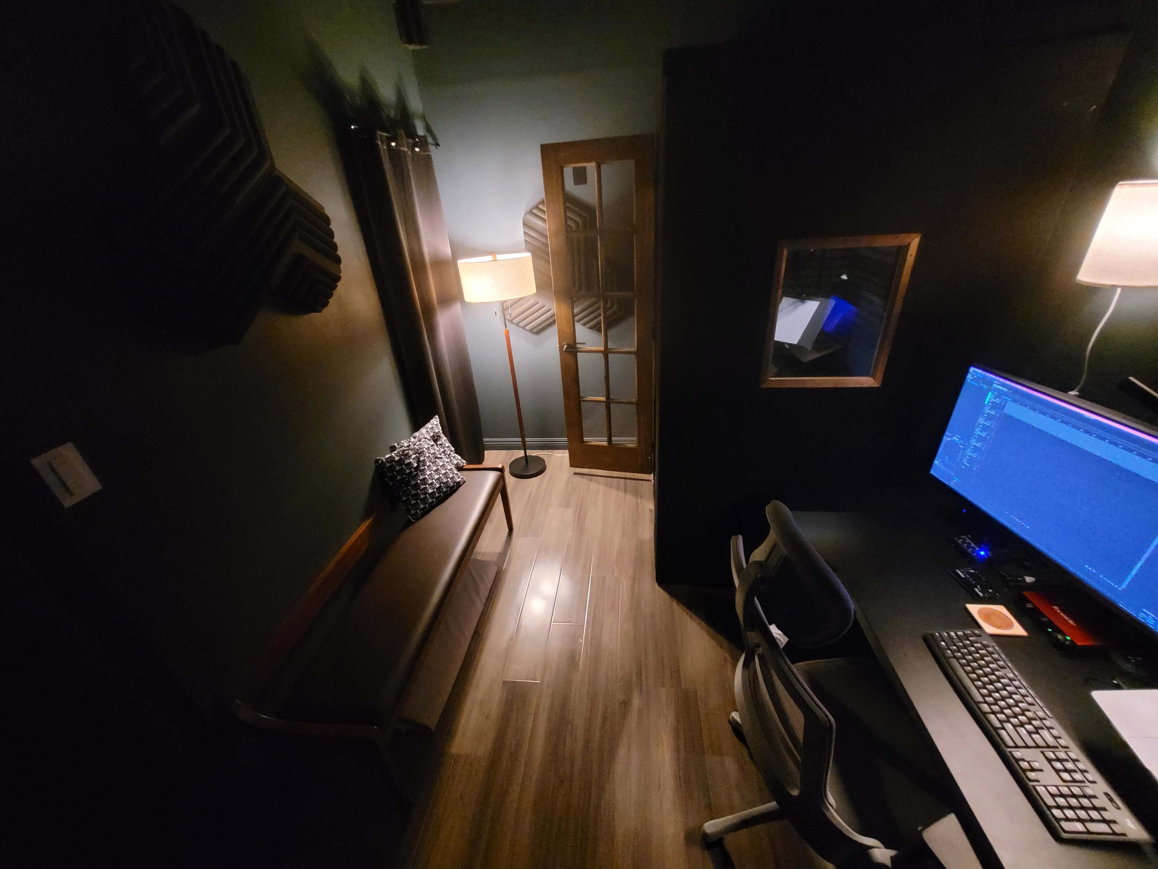 A dimly lit room featuring a desk with a computer, a sofa with a decorative pillow, and a lamp next to a glass-paneled door.