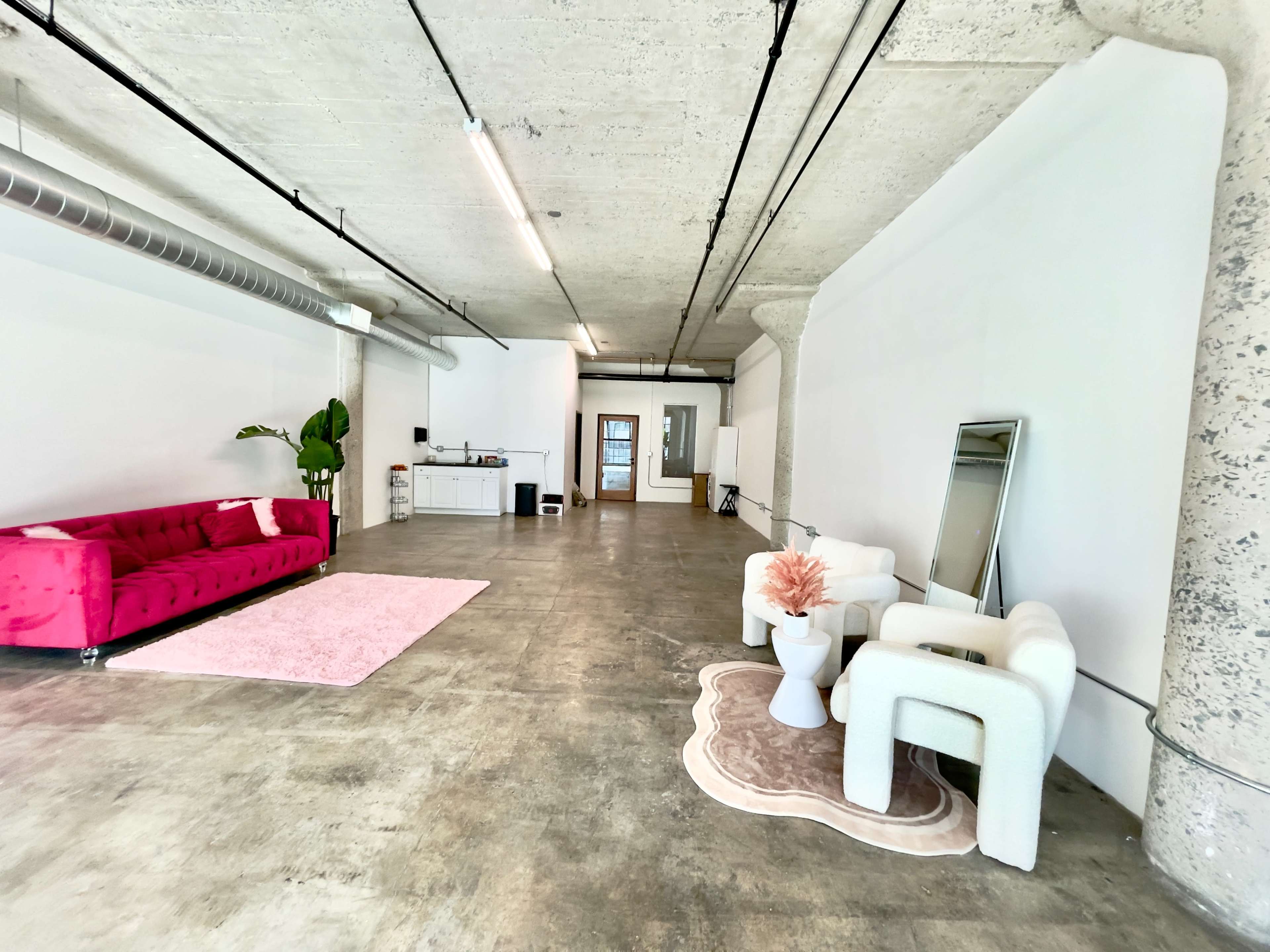 The image shows a spacious, minimalist interior featuring a pink couch, a white rug, and a small plant, with a kitchenette area in the background.