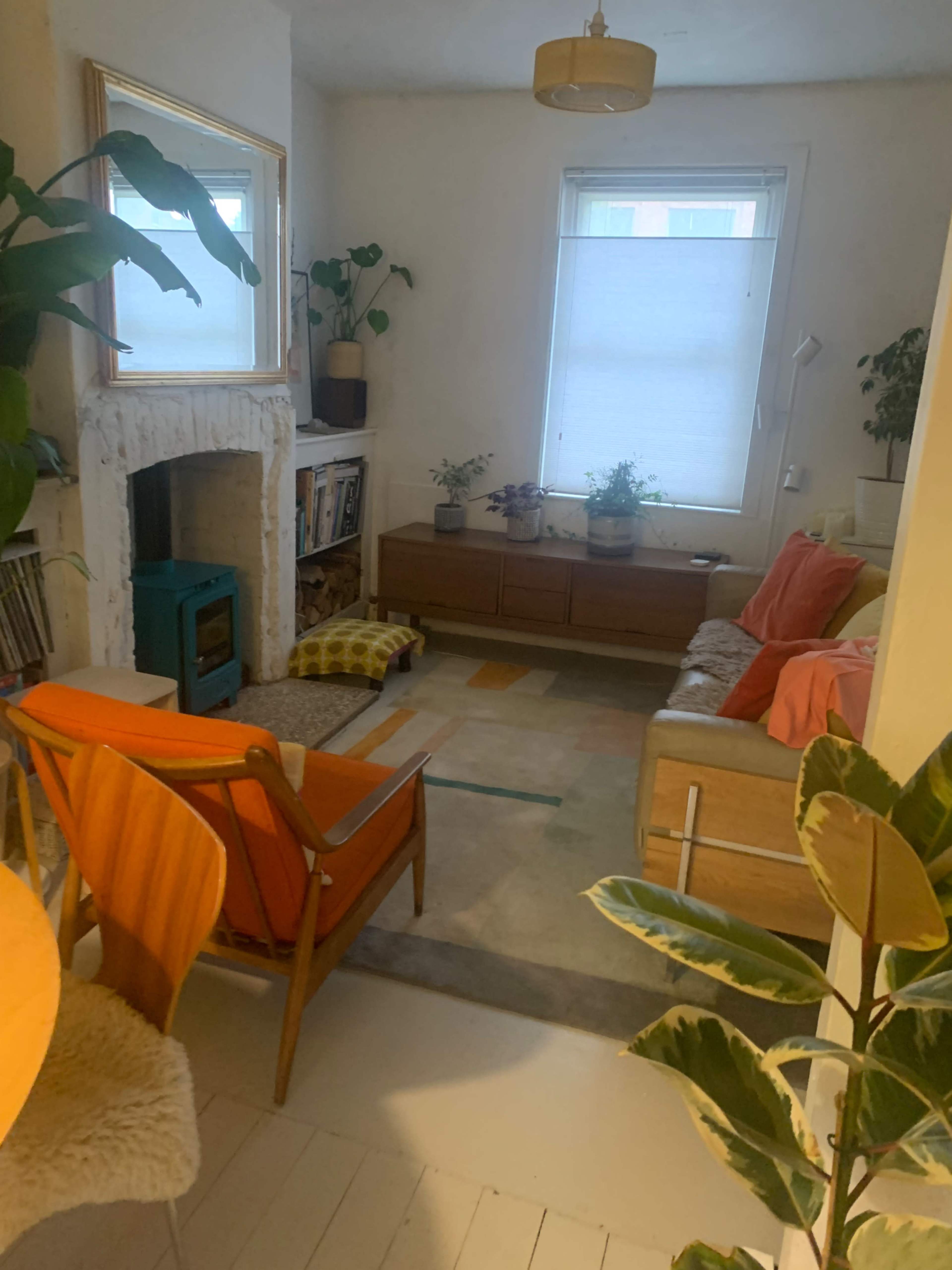 The image shows a cozy living room with a beige sofa, colorful chairs, a fireplace, plants, and a large window letting in natural light.