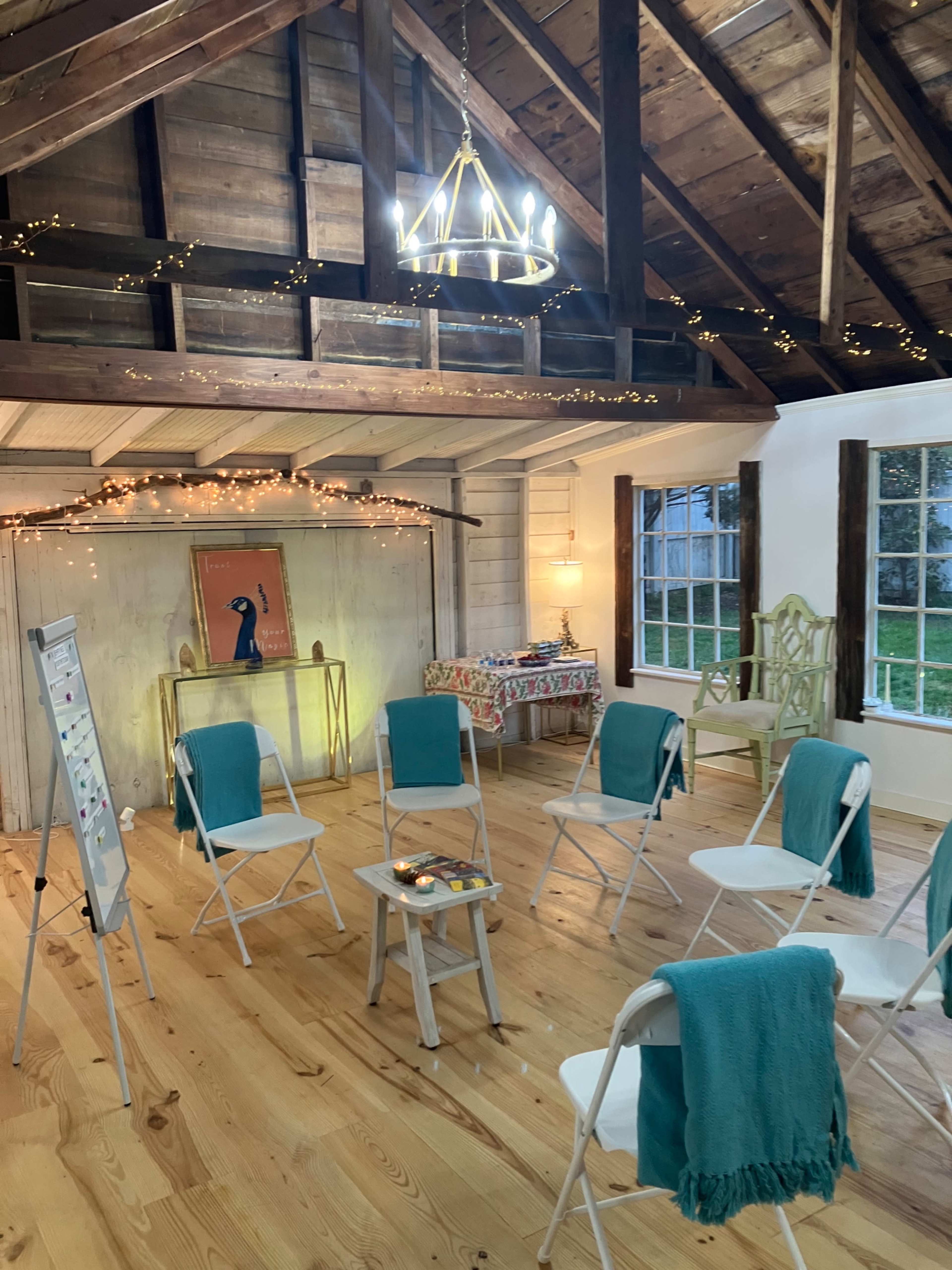 The room features a circle of white chairs with teal covers, surrounded by wooden beams, warm lighting, and decorative touches on the walls.