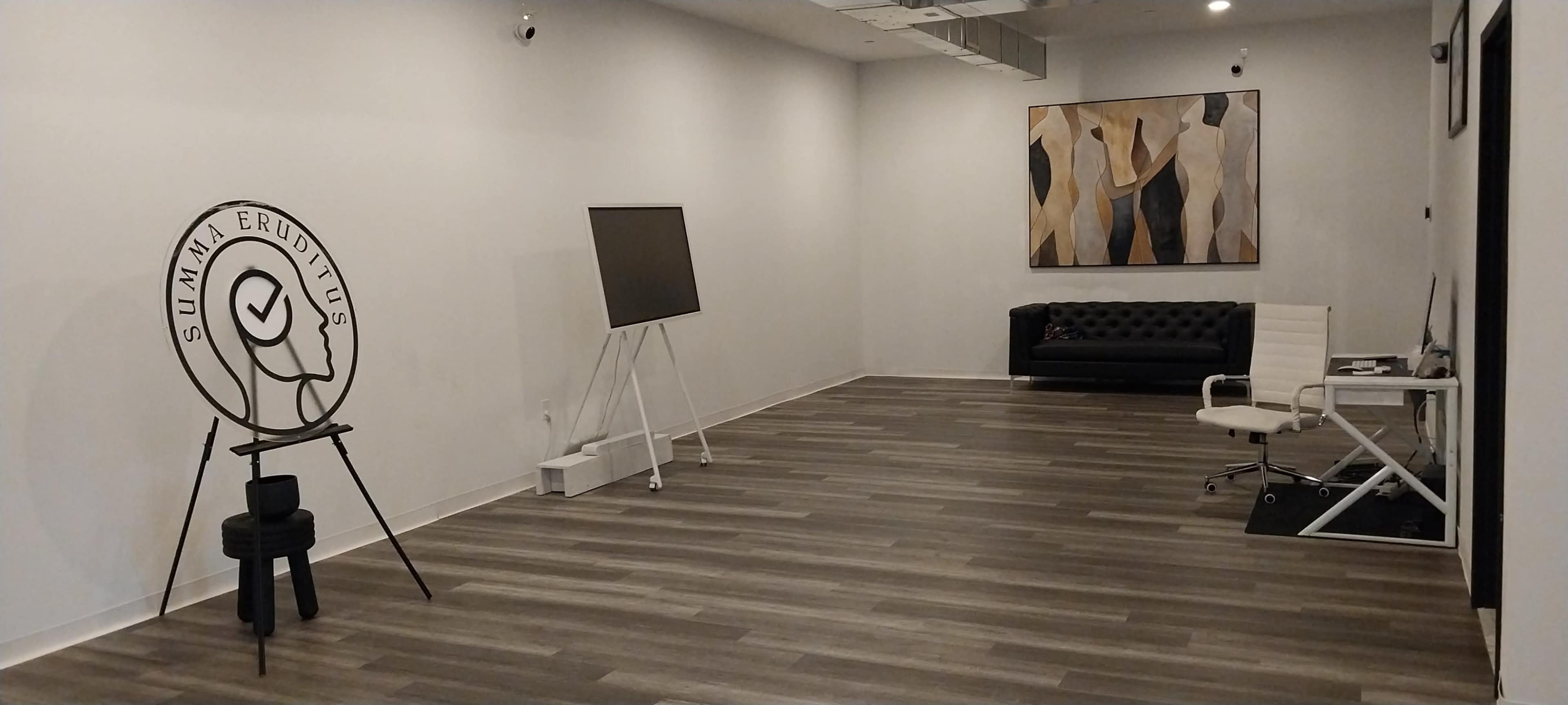 The image shows a spacious, minimalist room featuring a black sofa, a white chair and desk, an easel with a blank canvas, and a large abstract painting on the wall.
