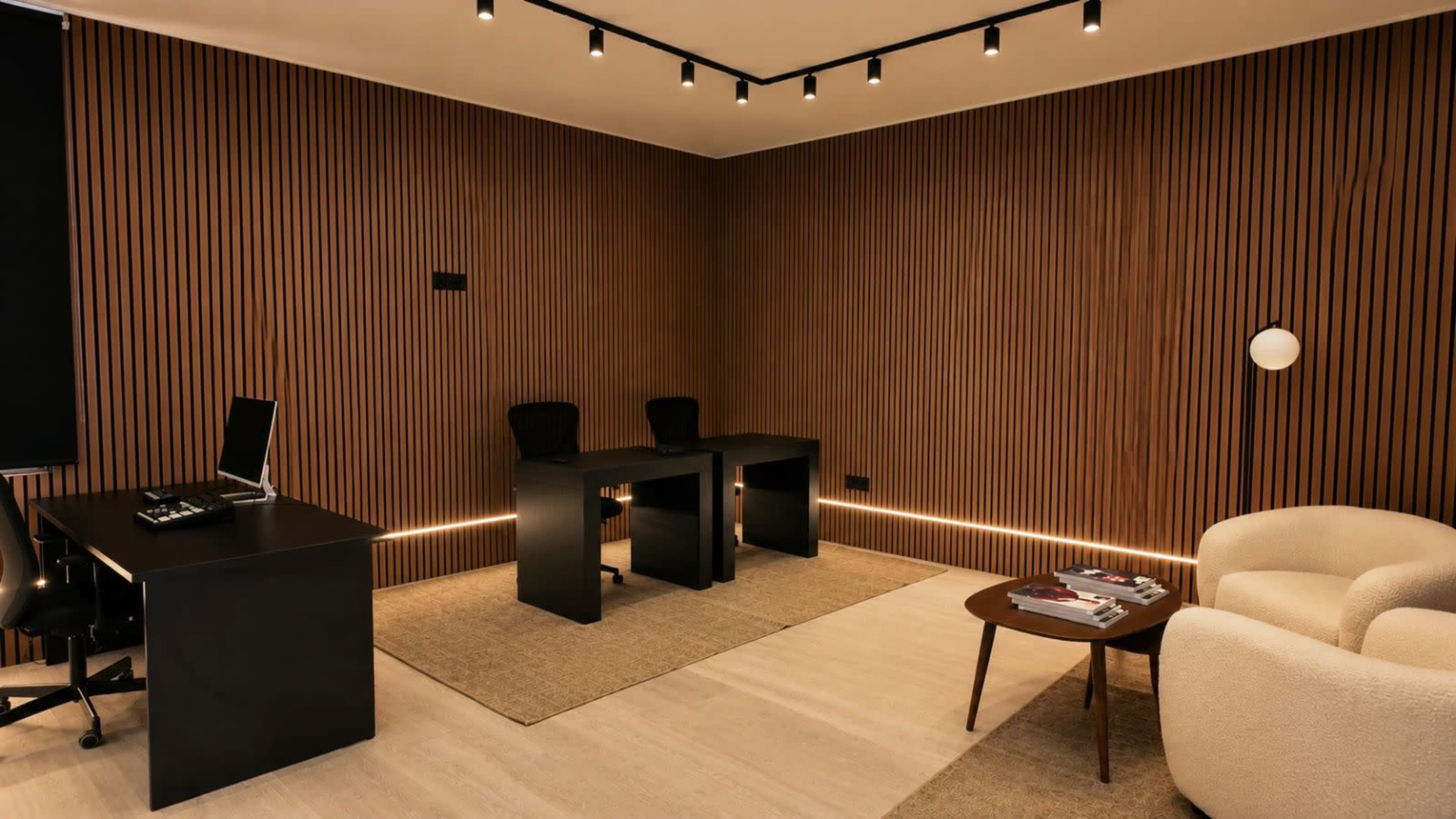 The image shows a minimalist office space with two black desks, matching chairs, and a round coffee table, all set against wooden paneled walls and soft lighting.