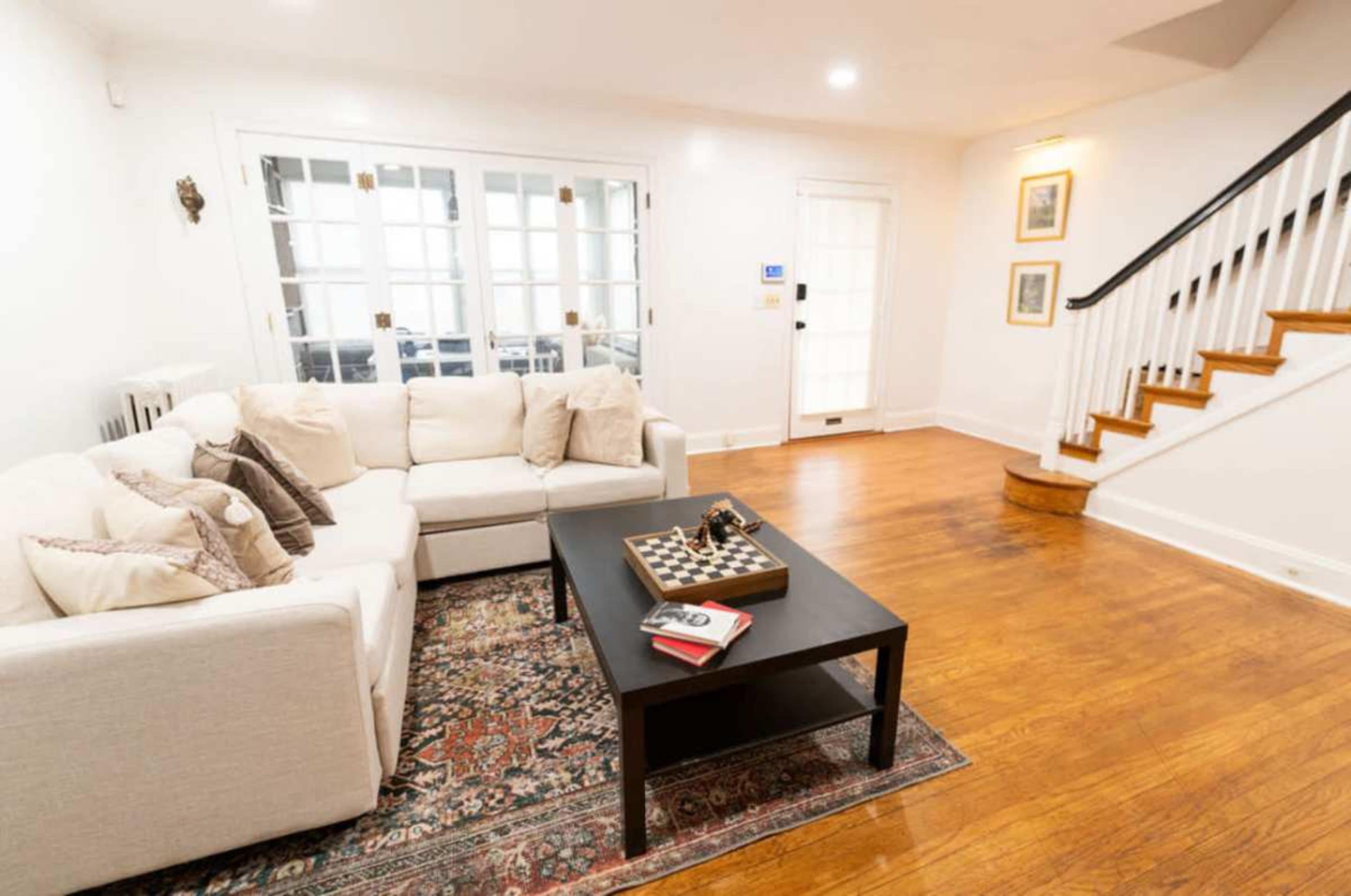 The image shows a bright living room with a sectional sofa, a coffee table featuring a chessboard, and a staircase leading to the upper level.