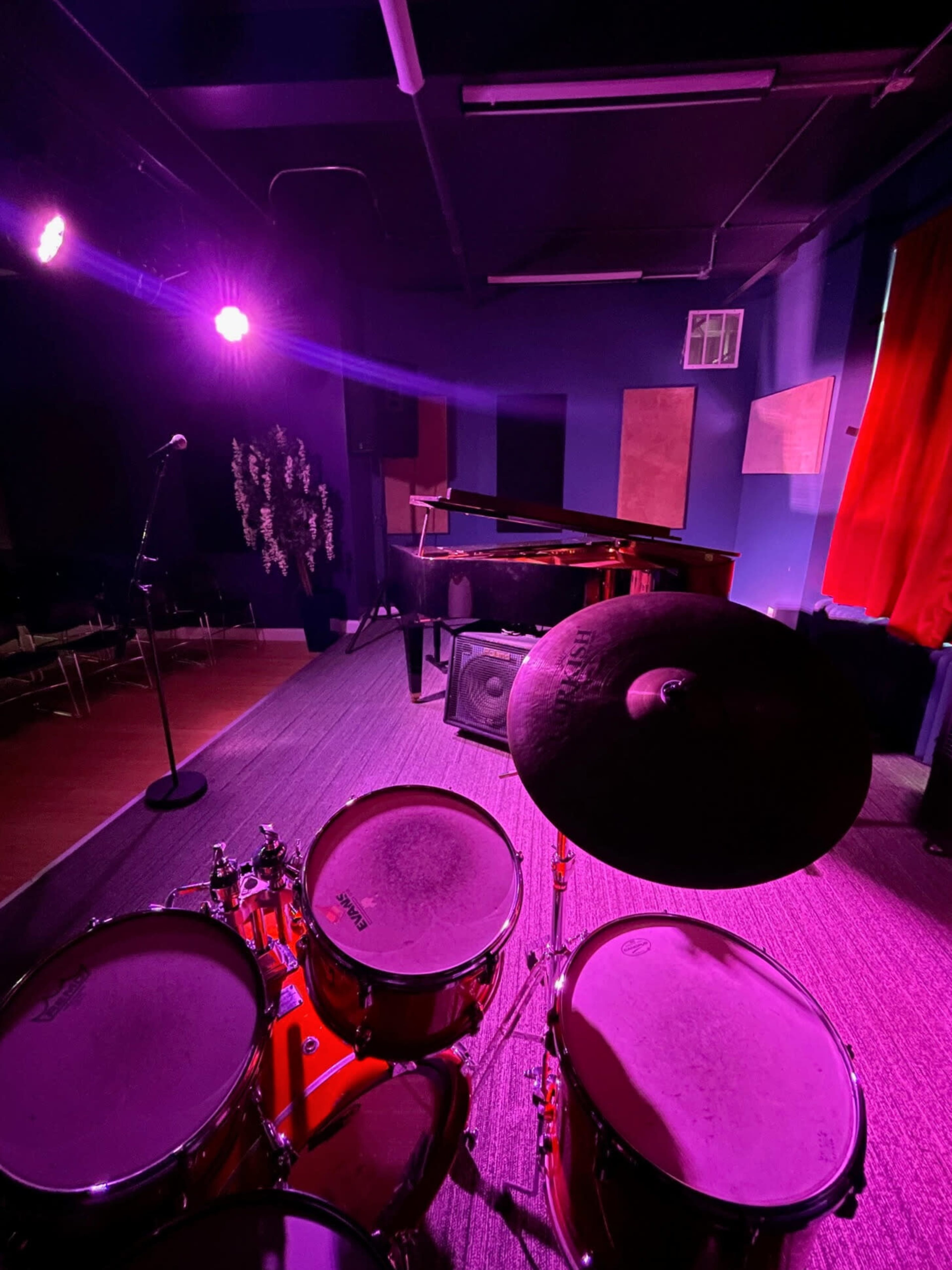 The image shows a small performance space with a drum set in the foreground, a piano in the background, and purple stage lighting.