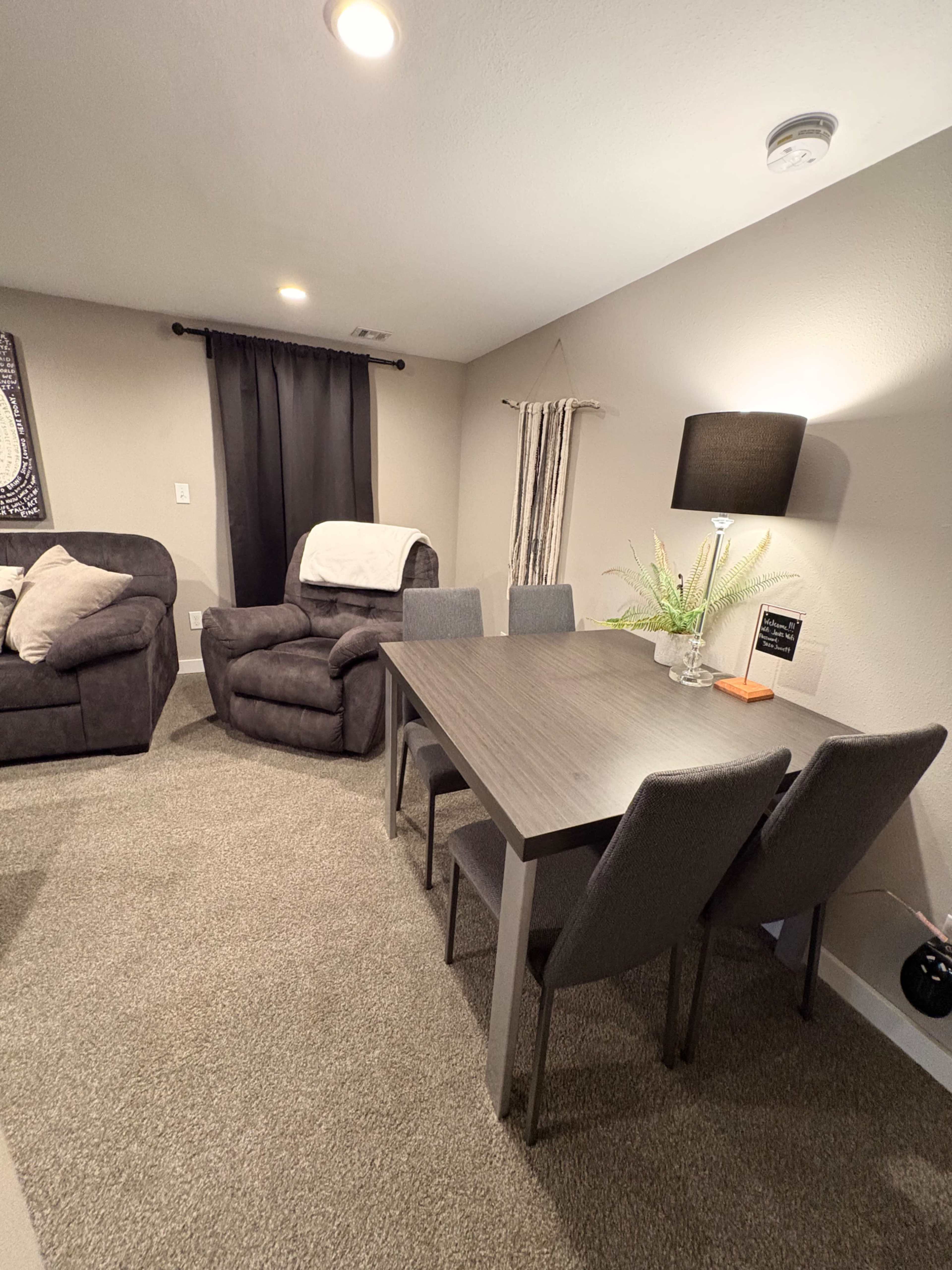 The image shows a cozy dining area with a rectangular table and four chairs, set against a backdrop of a living space featuring a recliner and soft furnishings.