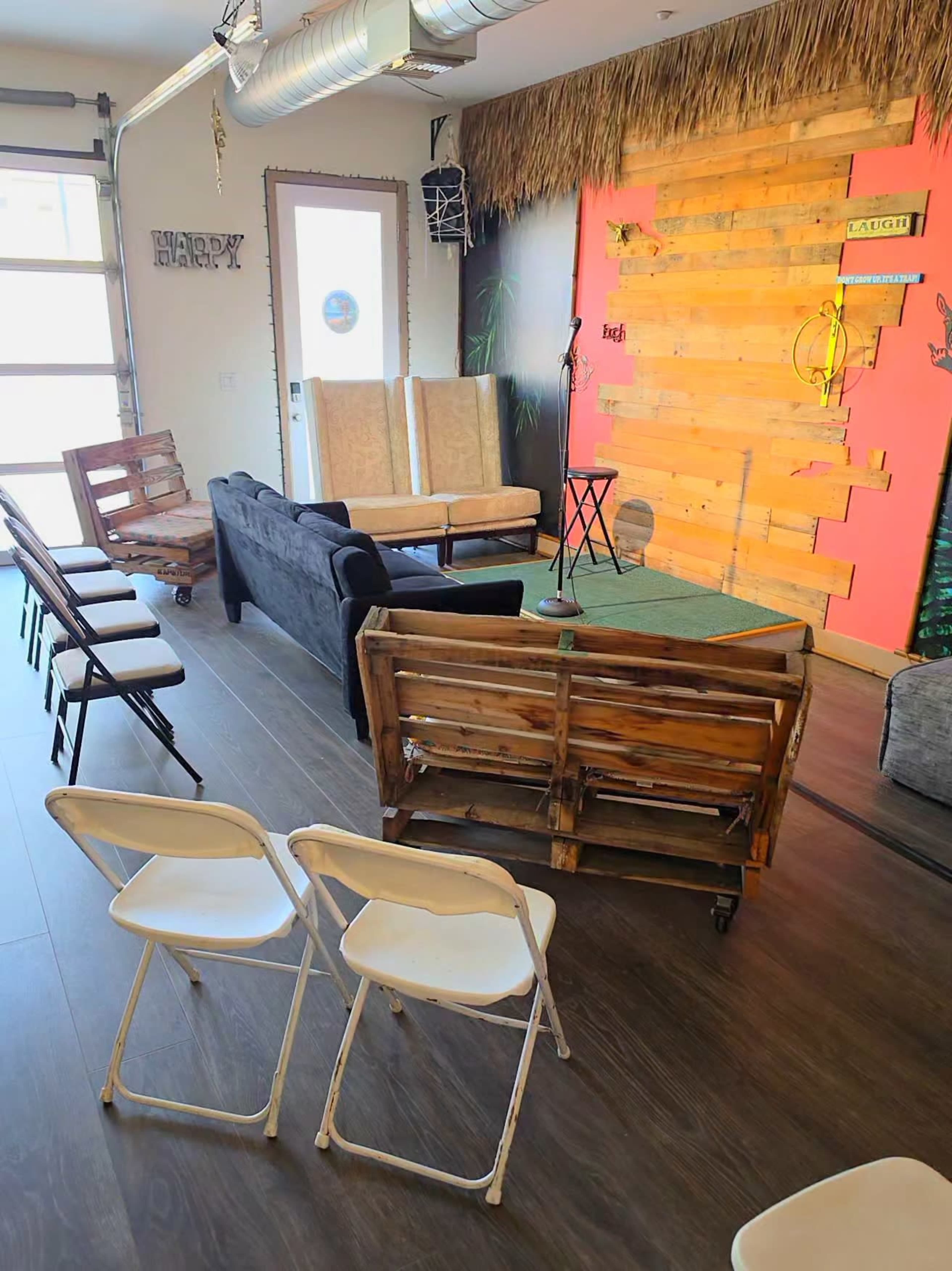 The image shows a room arranged for a performance, featuring various seating options, including wooden benches and folding chairs, facing a small stage with a microphone.