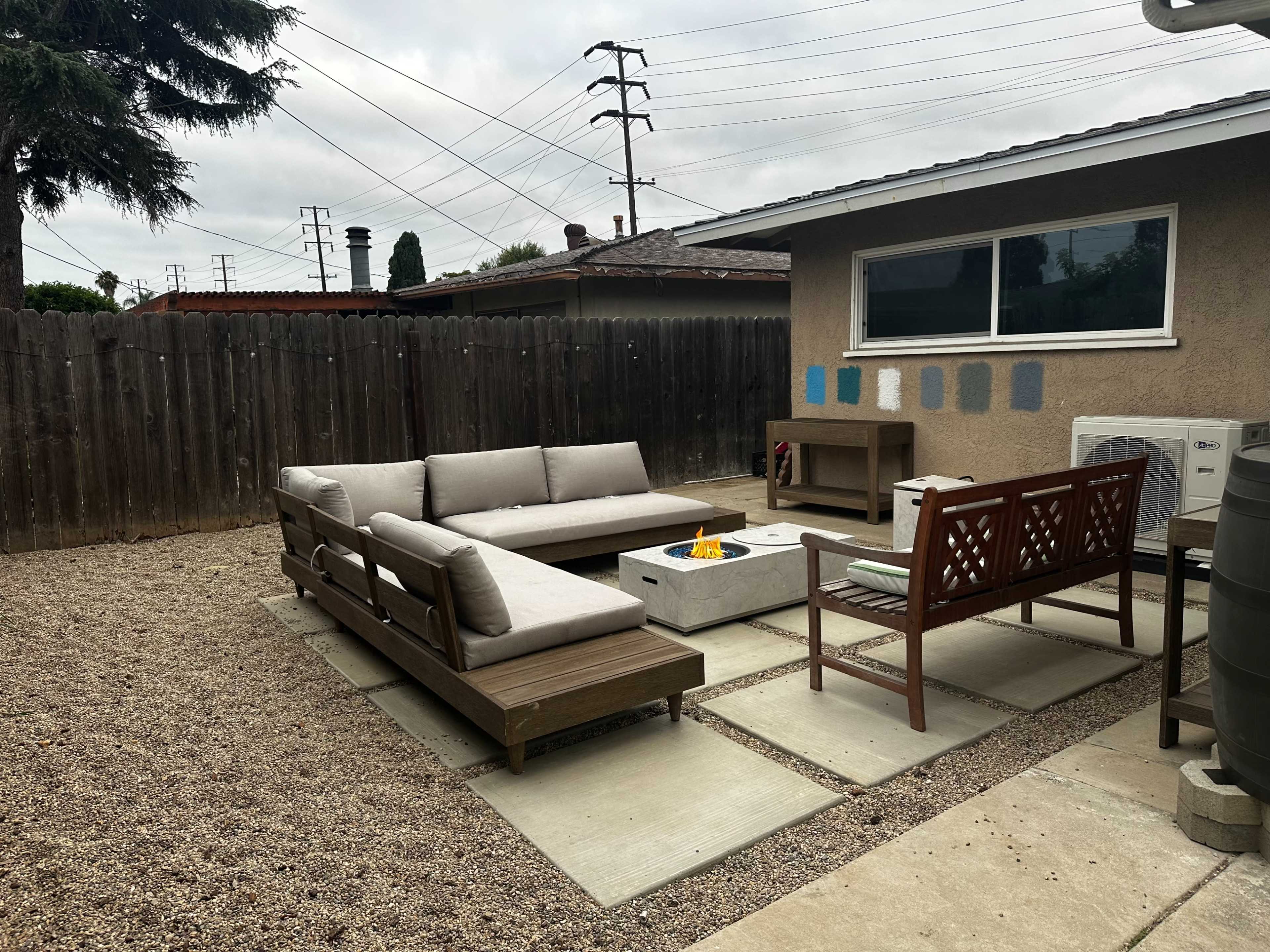 The image shows an outdoor seating area with a sectional sofa, a fire pit, and a wooden chair, surrounded by gravel and a wooden fence.