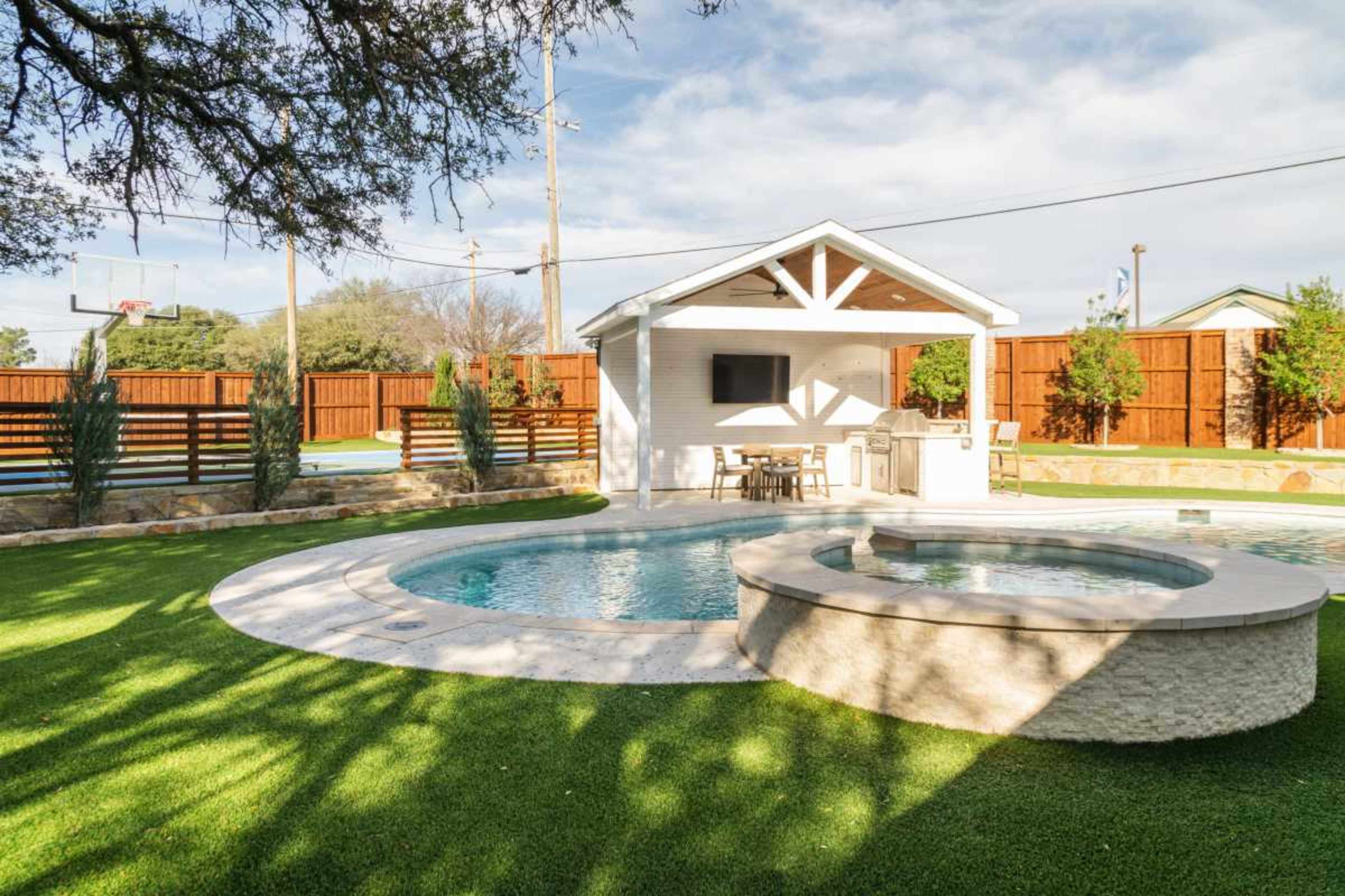 The image depicts an outdoor swimming area featuring a pool with an adjacent spa, surrounded by manicured grass and a small gazebo with seating and a television.