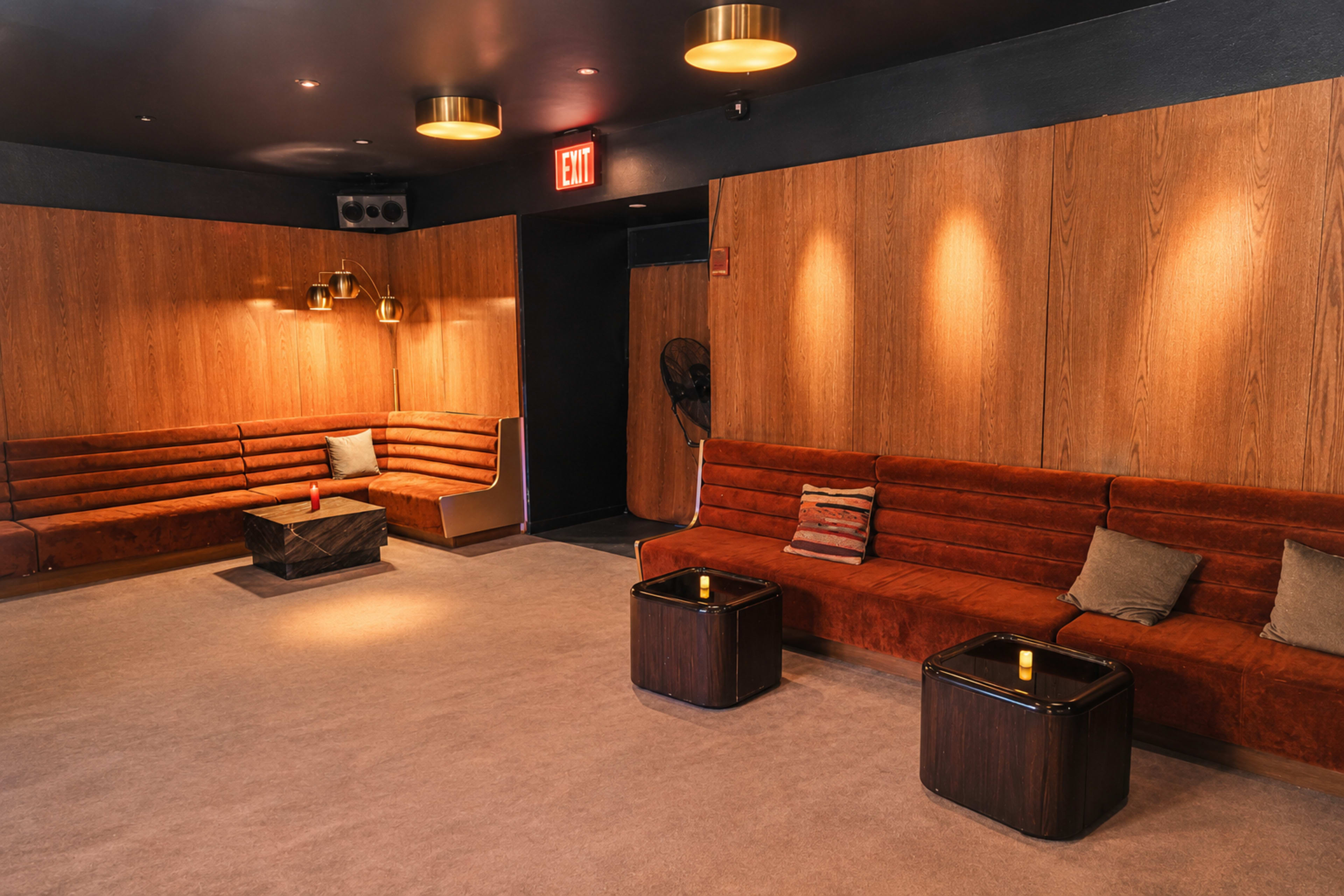The image shows a cozy lounge area with orange upholstered seating, wooden walls, and small, dark tables, illuminated by warm ceiling lights.