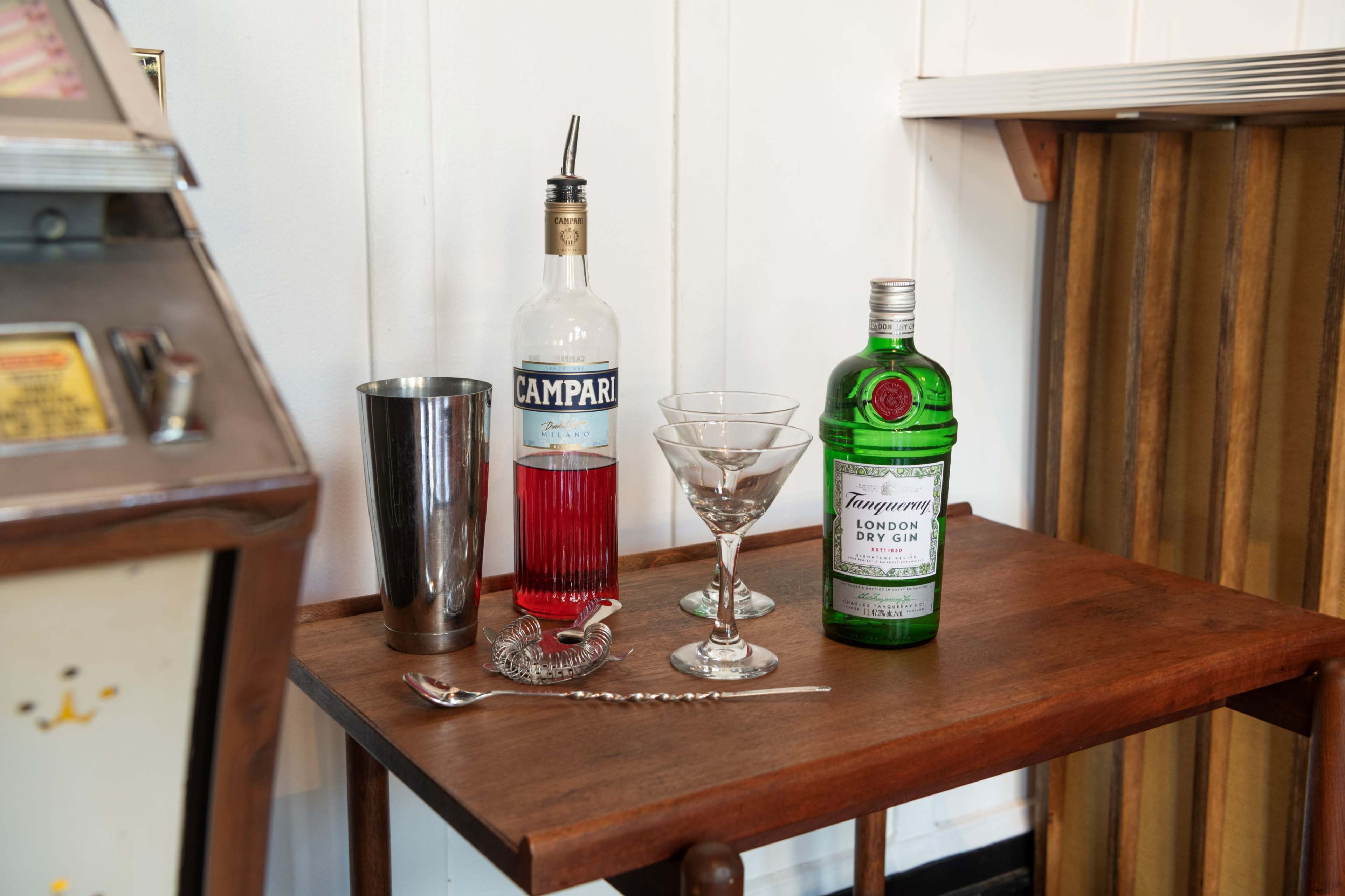 A wooden table displays a bottle of Campari, a bottle of Tanqueray gin, a cocktail shaker, and a martini glass.