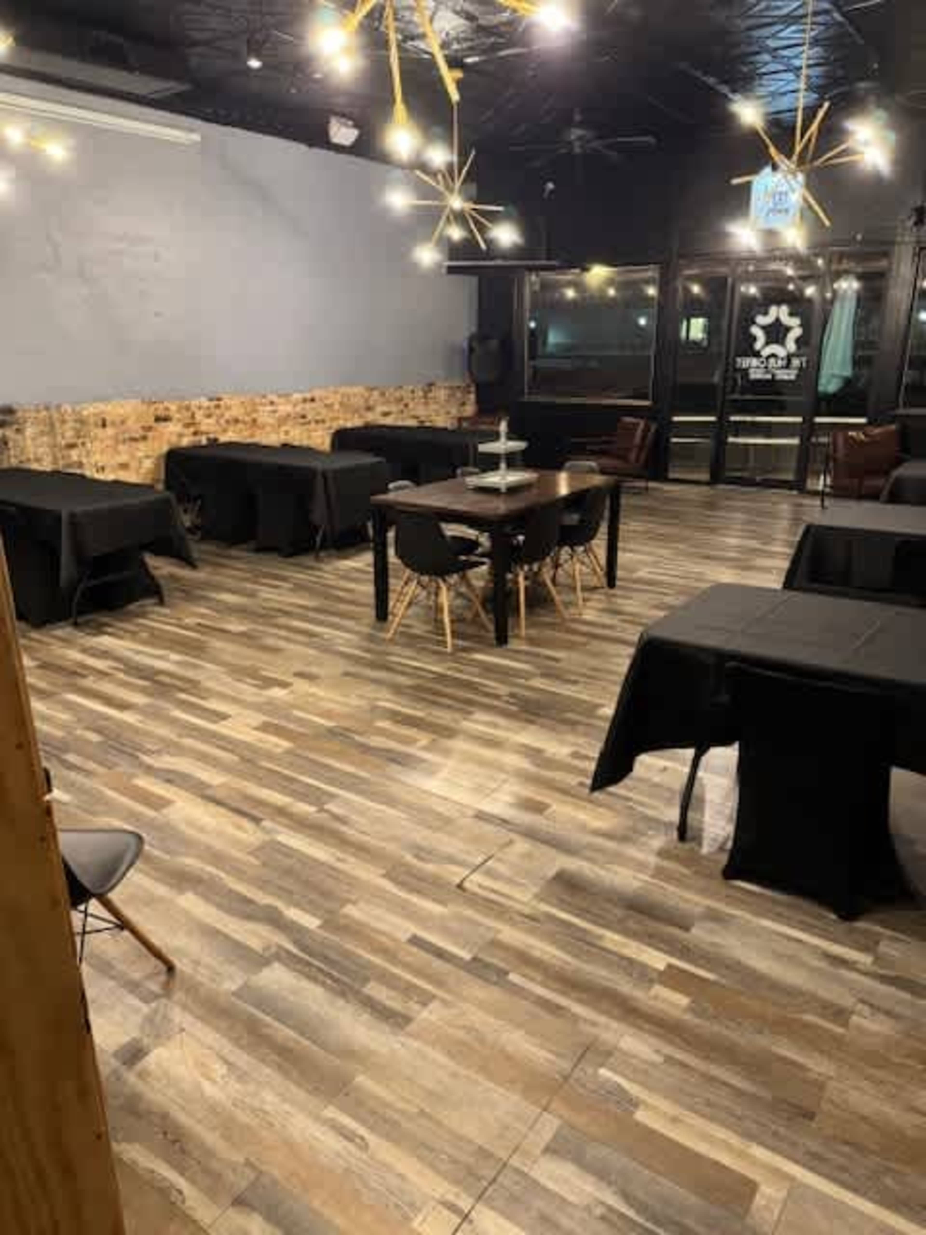 The image shows an interior of a restaurant or cafe featuring several black tables and chairs, wooden flooring, and modern lighting fixtures.