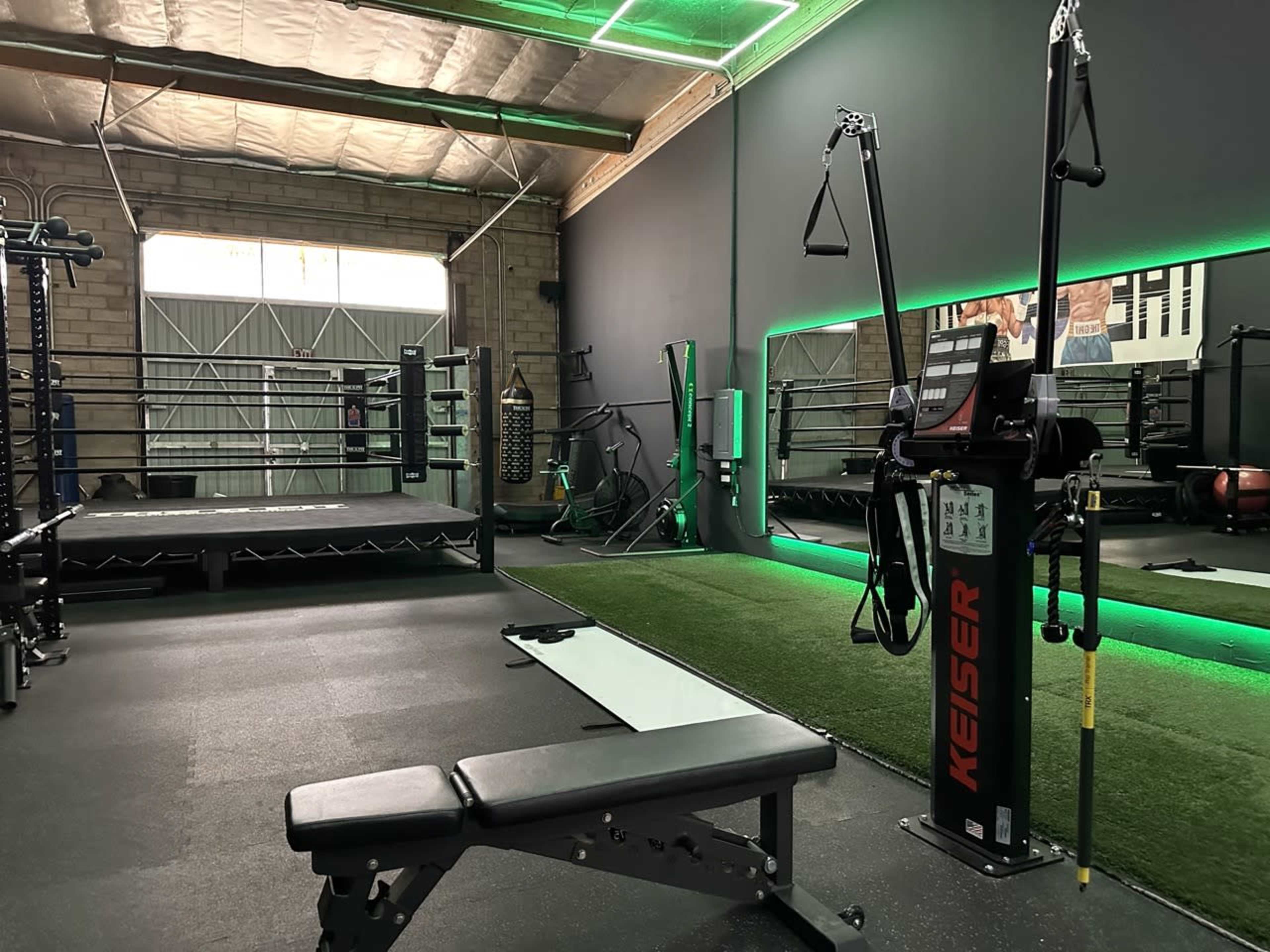 A modern gym interior featuring a weight bench, resistance training machine, boxing ring, and green turf area under bright lighting.