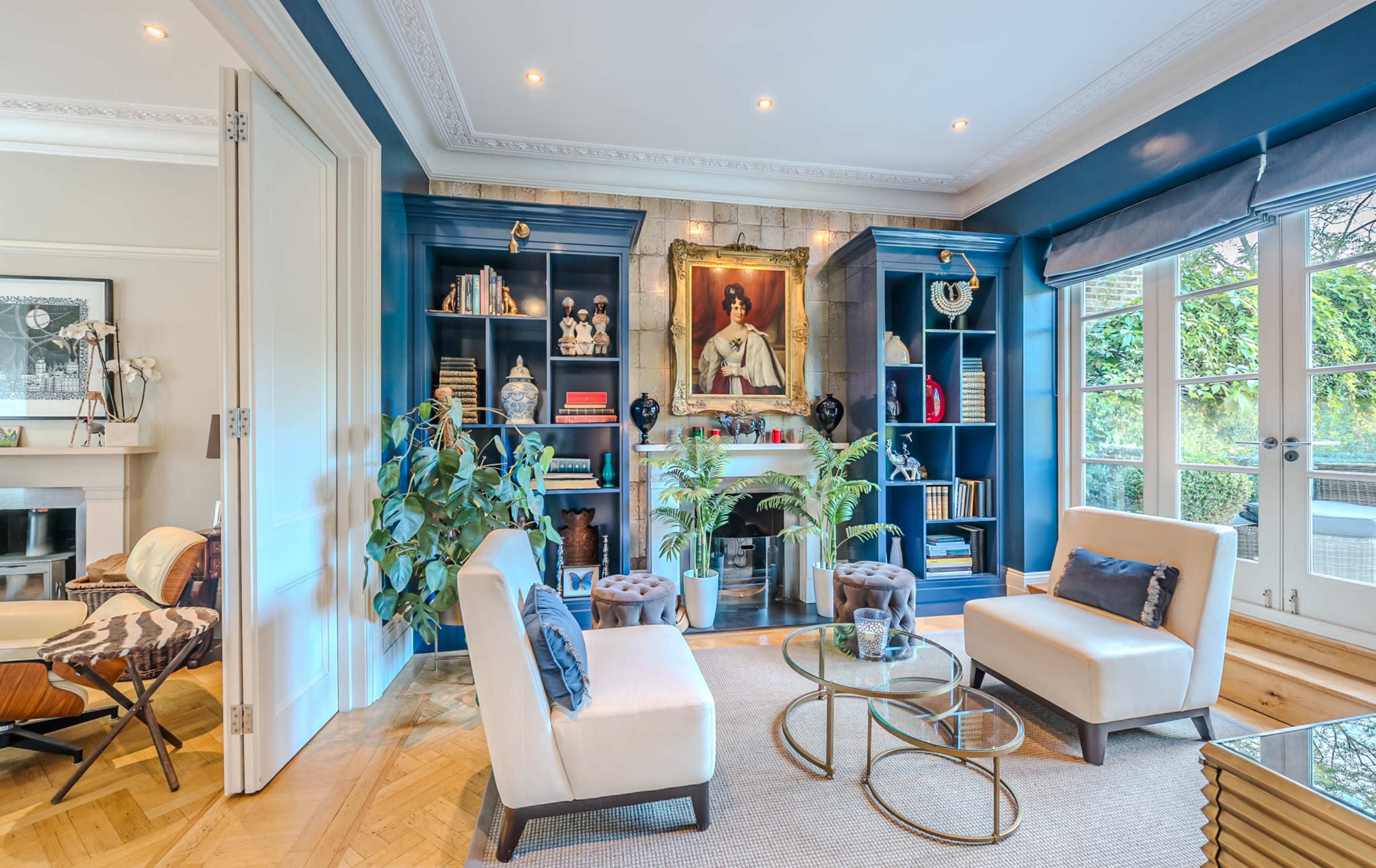 The room features two cream-colored chairs and a glass coffee table, with blue built-in shelving and a large portrait hanging above a fireplace.
