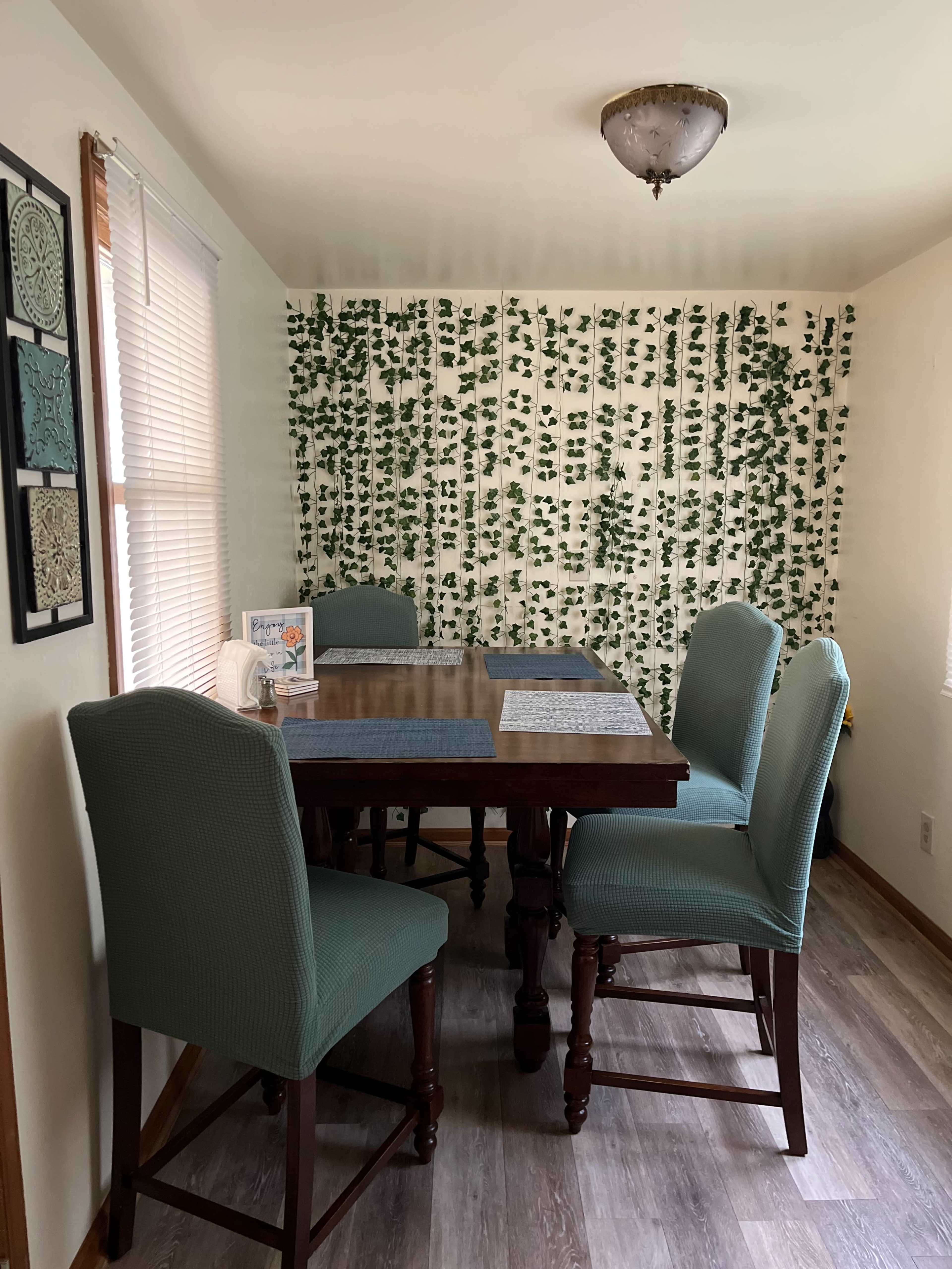 A dining area features a wooden table with four upholstered chairs, surrounded by a wall adorned with green vines.