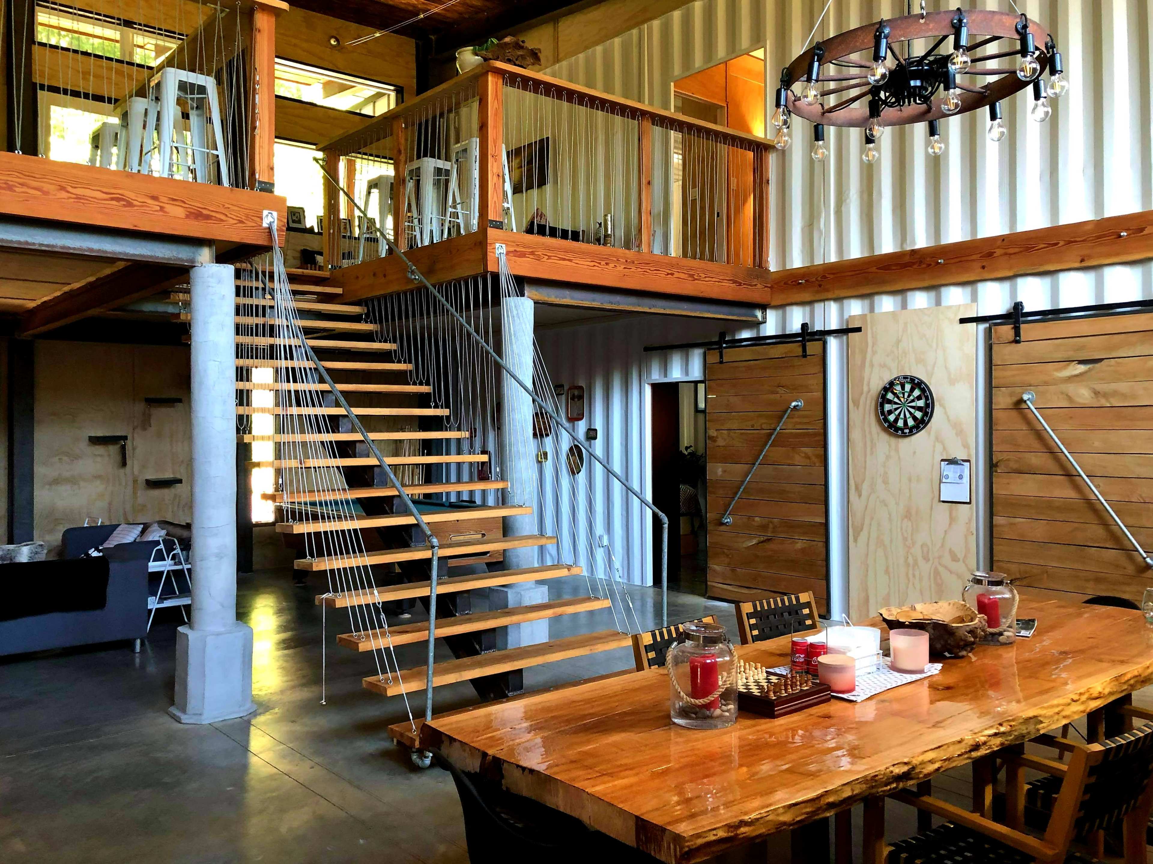 An industrial-style interior with a wooden dining table, a staircase leading to a loft area, and large metal beams framing the space.