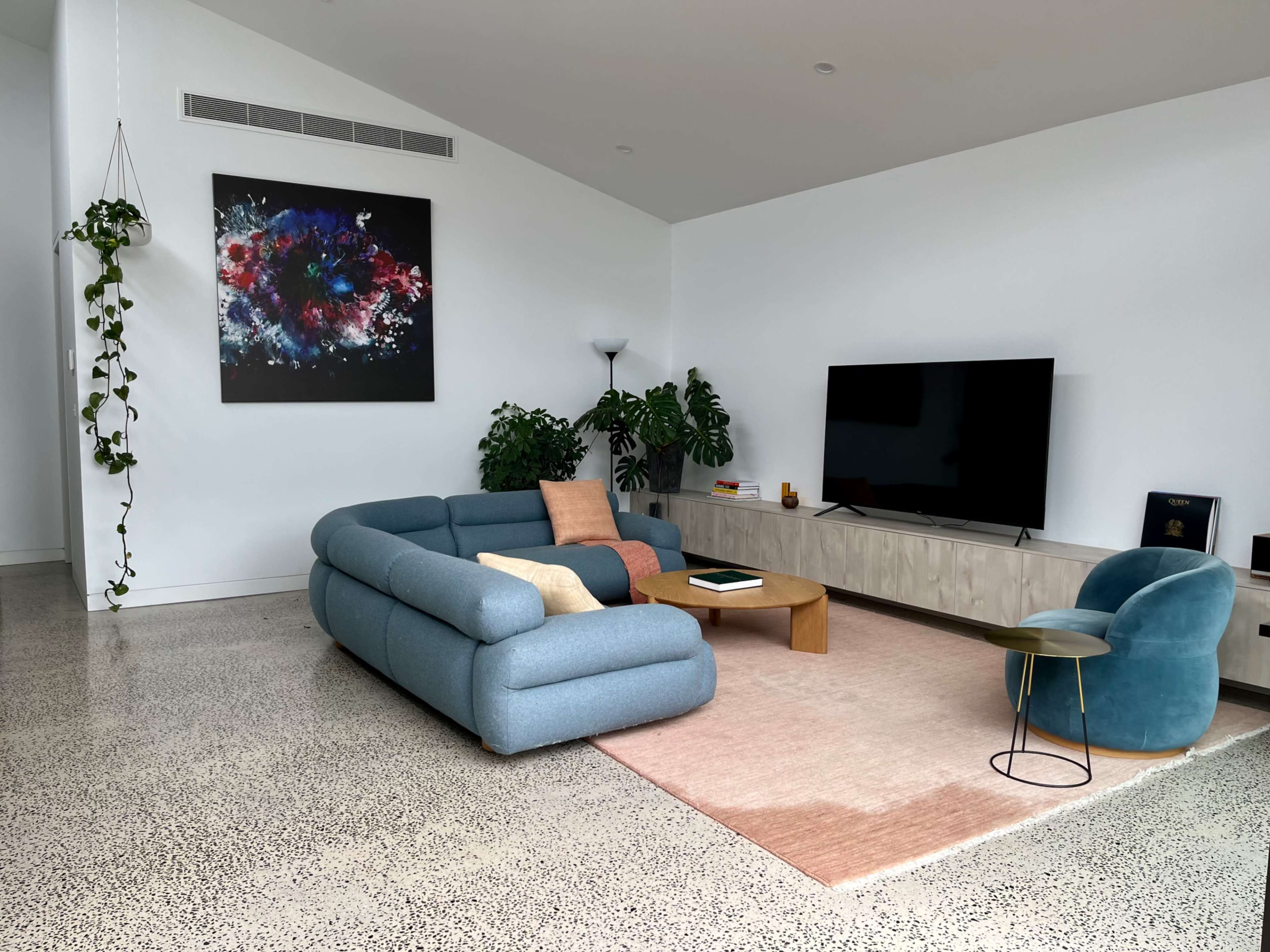 A modern living room features a blue curved sofa, a wooden coffee table, a television on a light-colored media unit, and green plants against white walls.
