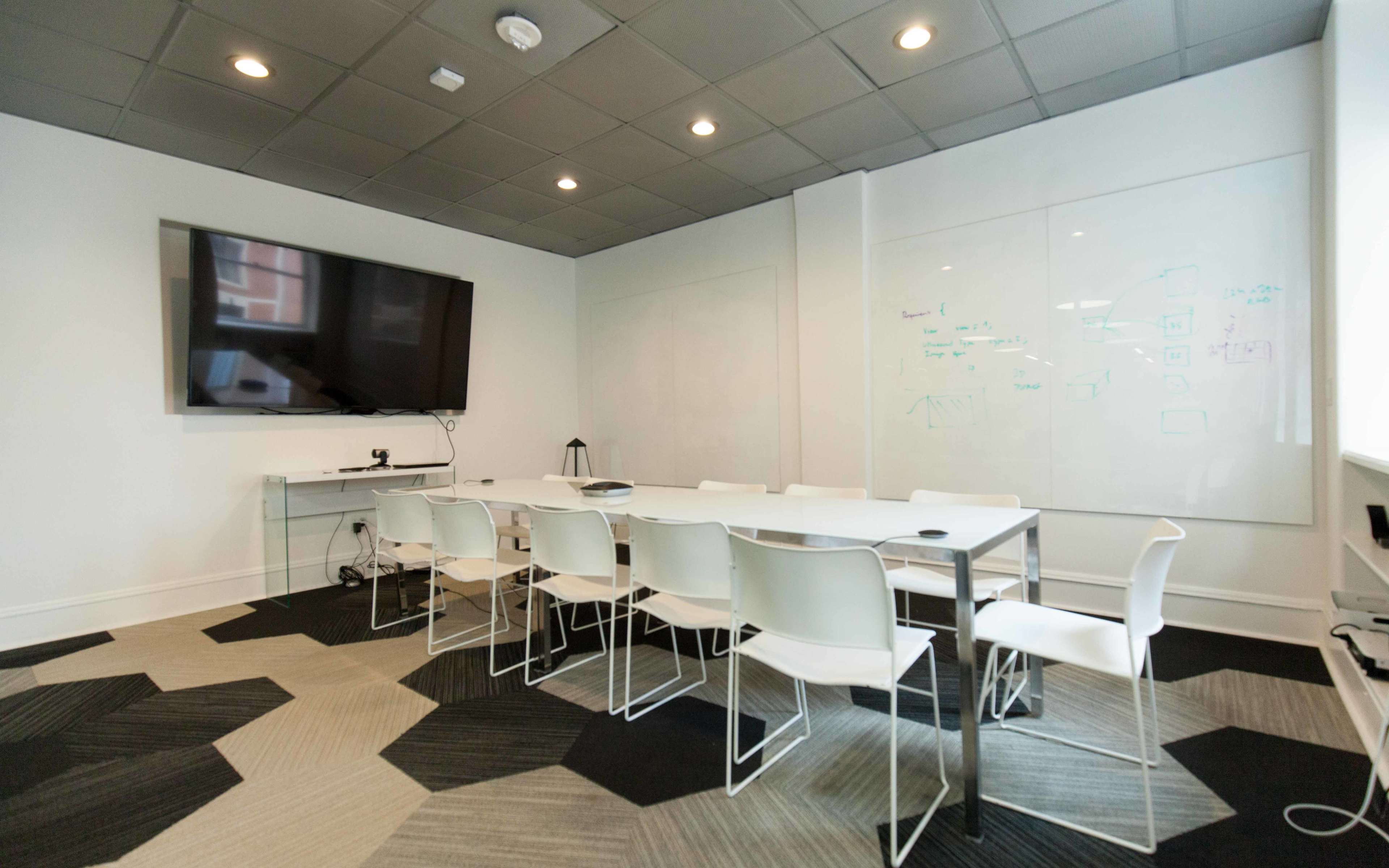 A modern conference room features a large TV screen, a whiteboard, and a long table surrounded by several white chairs on a patterned carpet.